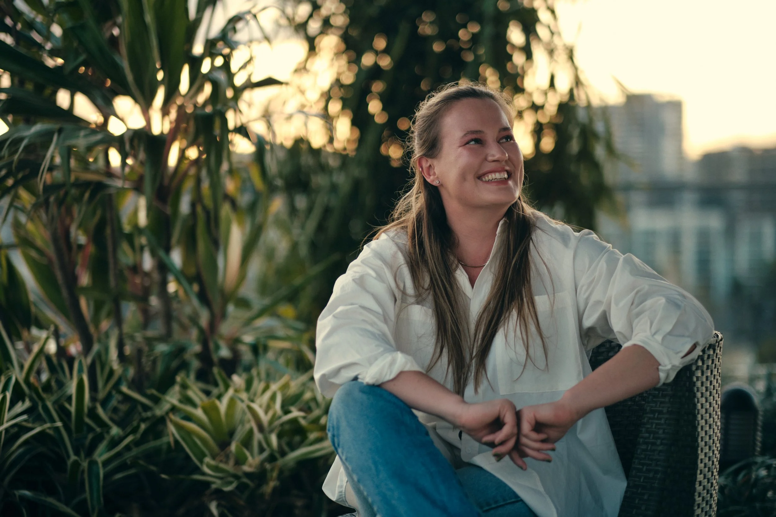 A young woman with long brown hair, wearing a white shirt and blue jeans, smiling while sitting outdoors in a garden or park during sunset. She is surrounded by green plants and trees, with a cityscape in the background.