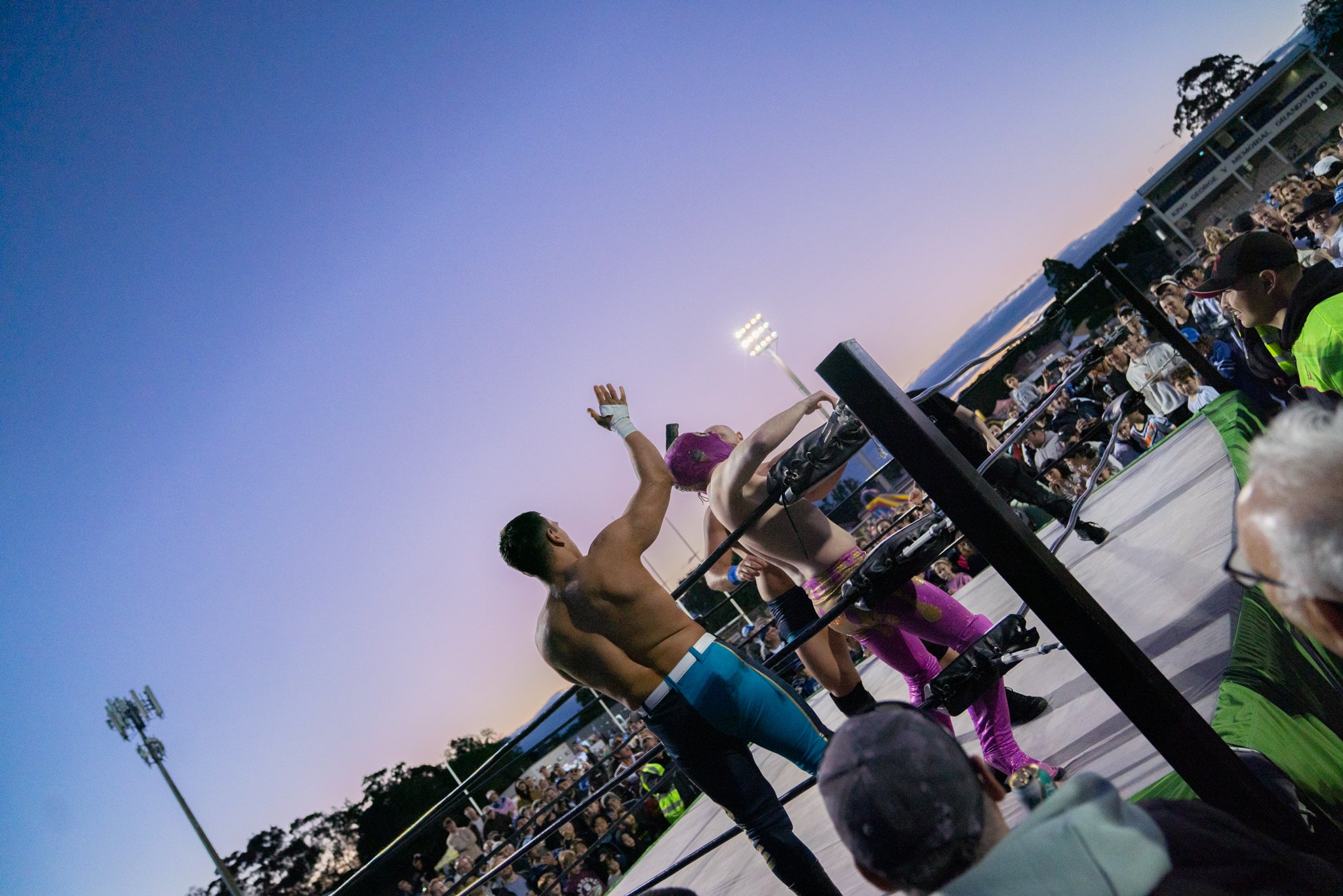 Wrestlers in colorful outfits greeting each other in a wrestling ring at sunset, surrounded by spectators.