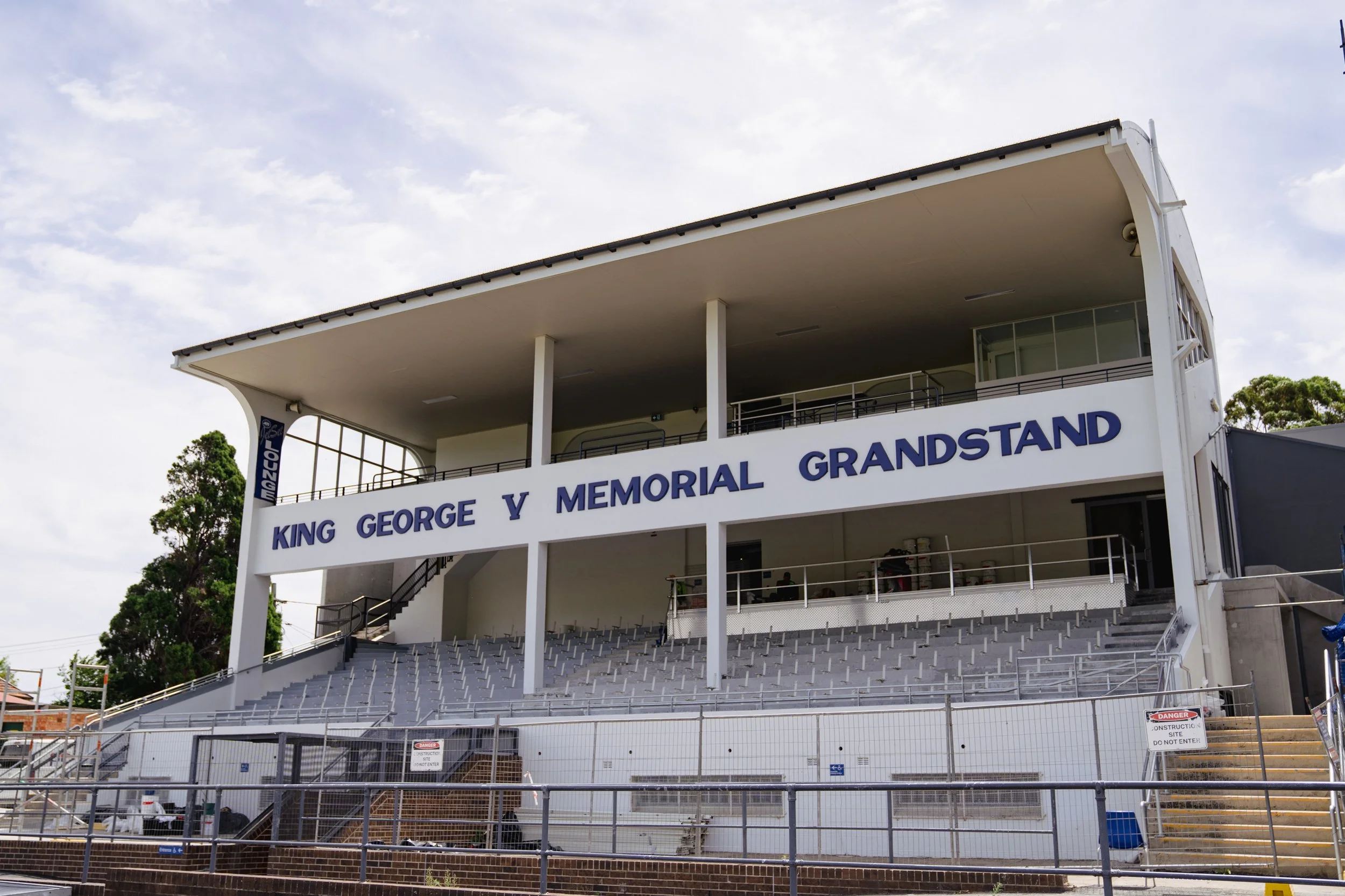 An empty outdoor grandstand with seating and a white building, labeled 'King George V Memorial Grandstand,' with a sign indicating construction and danger, under a partly cloudy sky.