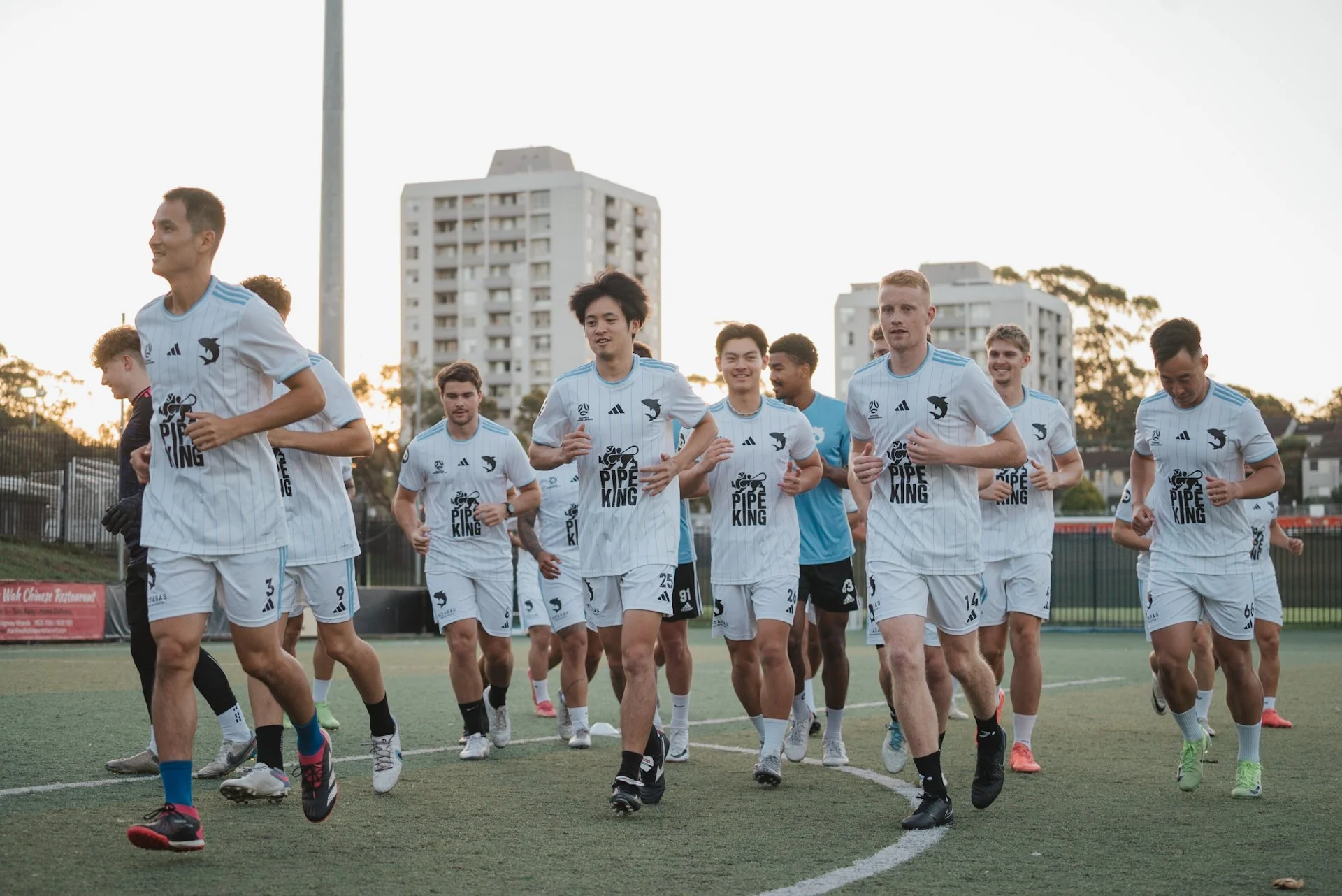 A group of soccer players jogging on a field during sunset, wearing white jerseys with blue stripes and a black logo that says 'Pipe King'.