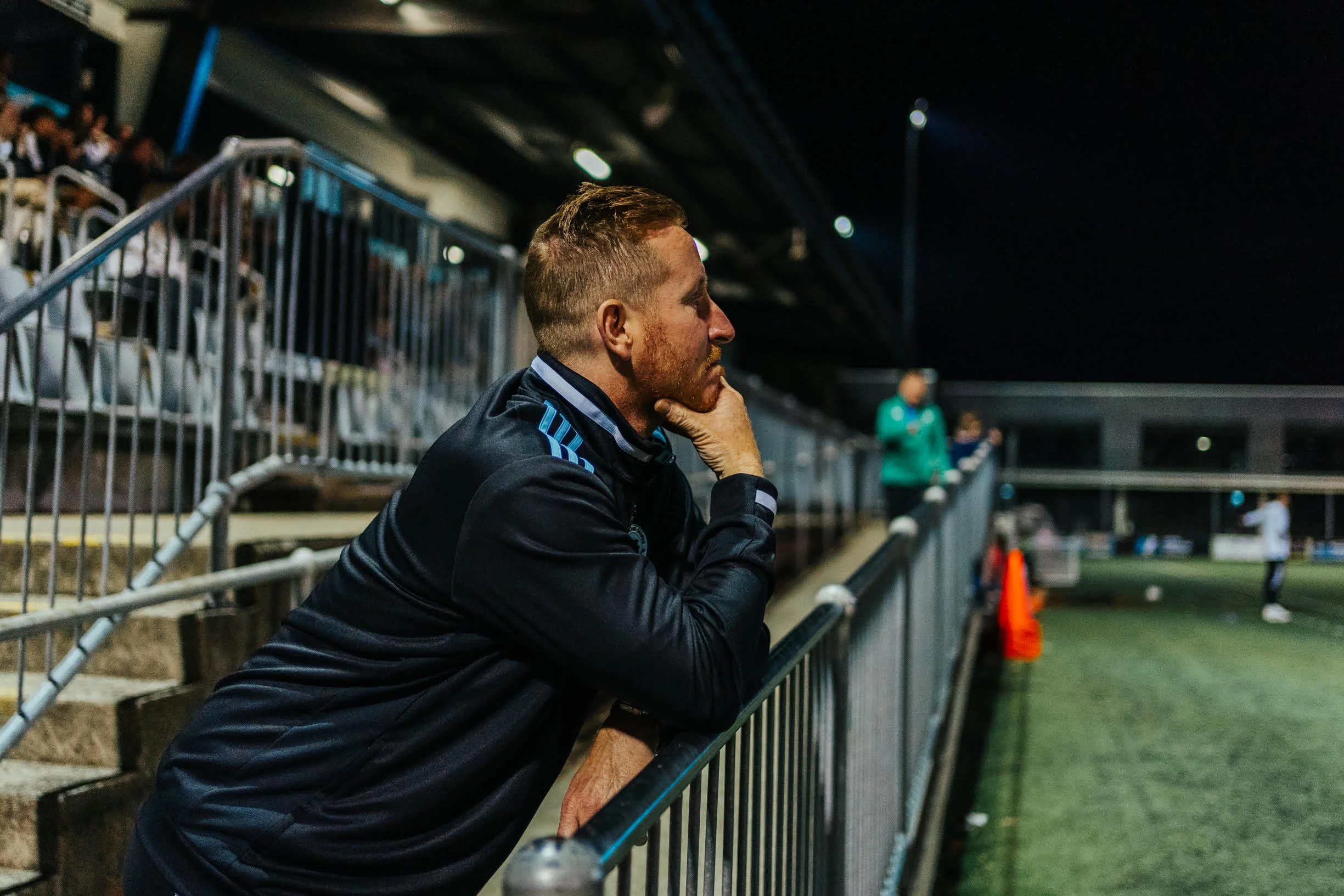 A man watching a sports game at night, leaning on a railing and resting his chin on his hand, with a stadium and other spectators in the background.