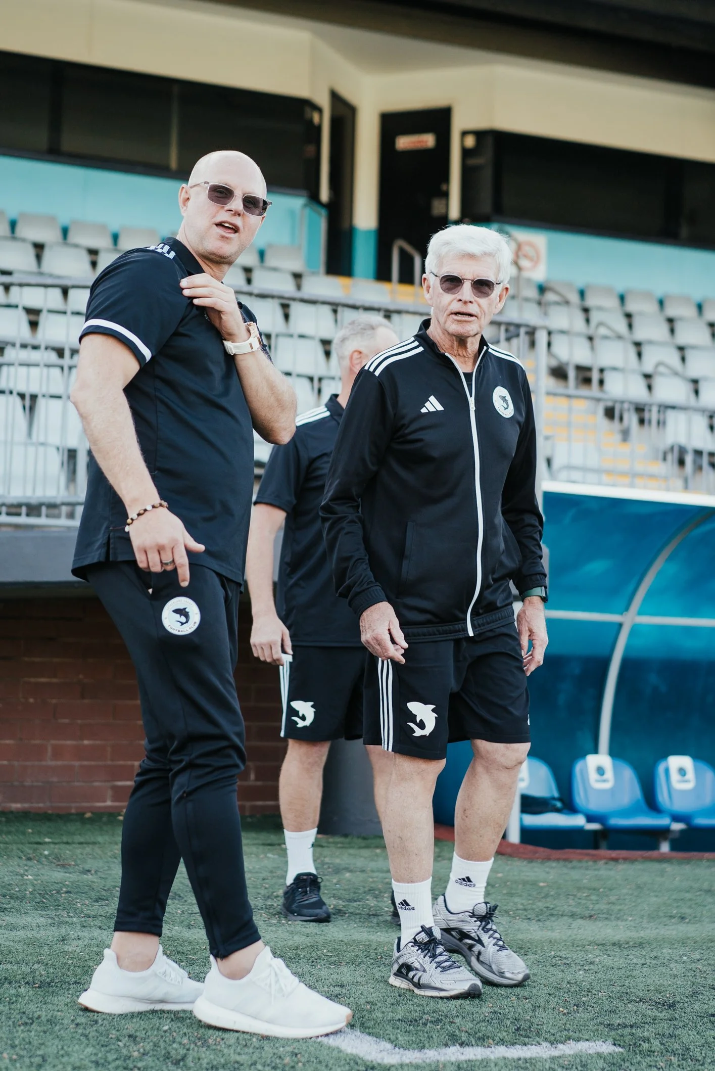 Two men in sportswear standing on a sports field, wearing sunglasses and black Nike outfits with a dolphin logo, with empty bleacher seats in the background.