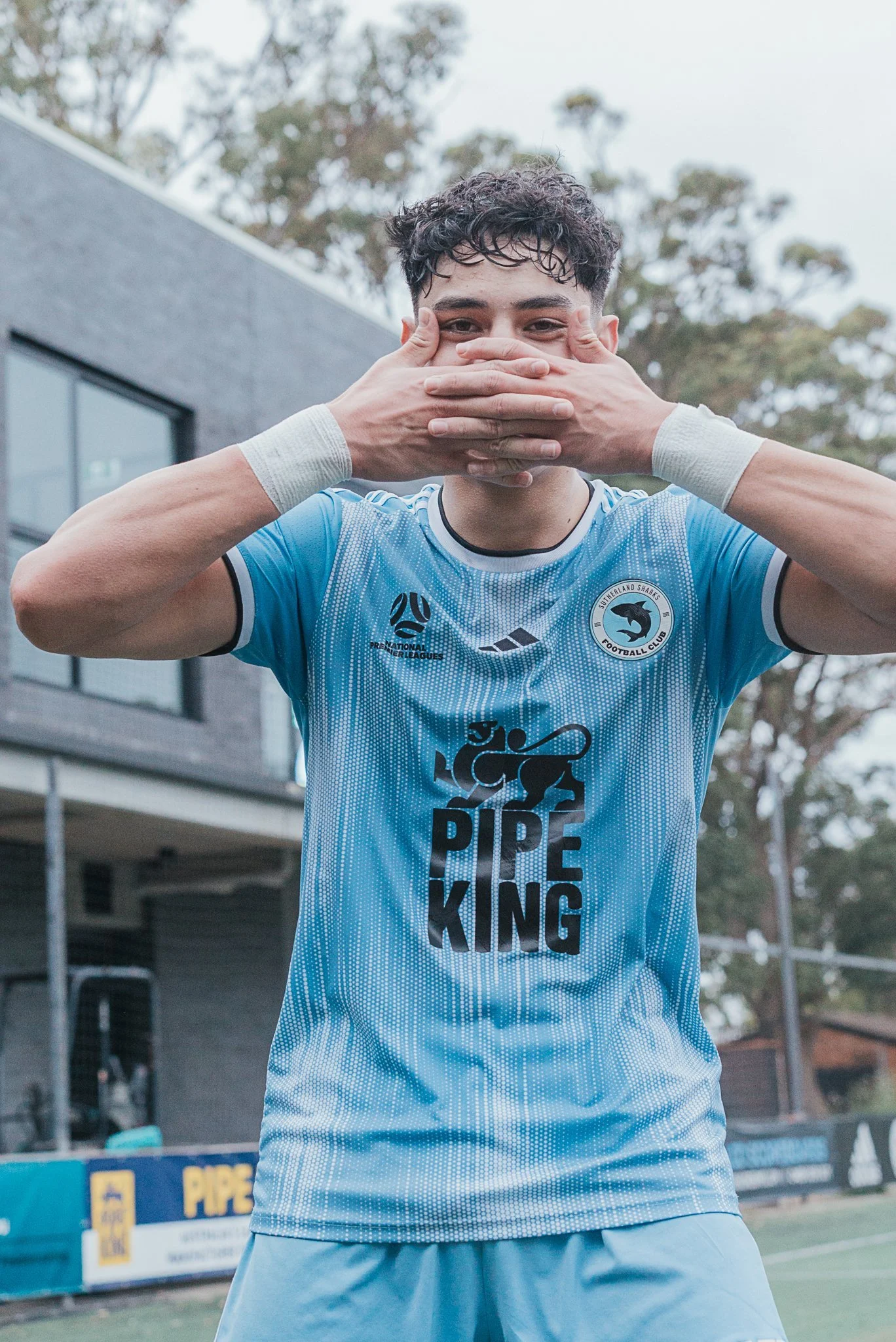 A young male soccer player in a light blue jersey standing outdoors on a soccer field, covering his mouth with his hands, with a building and trees in the background.