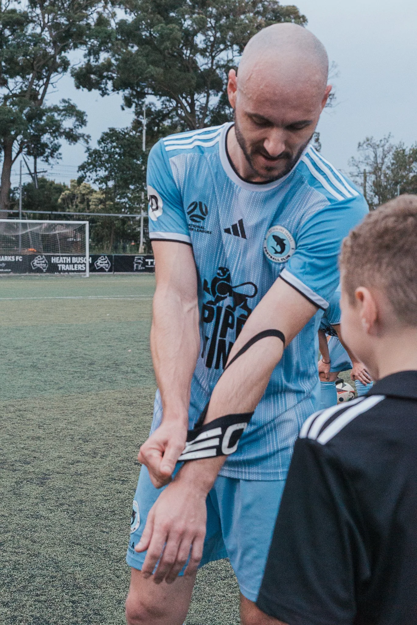 A man in a blue soccer jersey is helping a young boy in a black athletic top adjust his sleeve on a soccer field. They are surrounded by trees and stadium equipment.