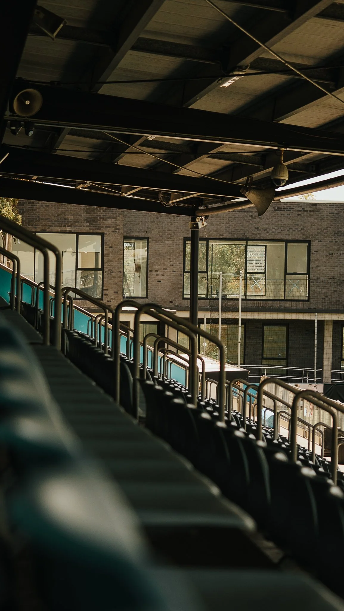 Empty sports stadium seating with green chairs, metal railings, and a view of a brick building with large windows in the background.