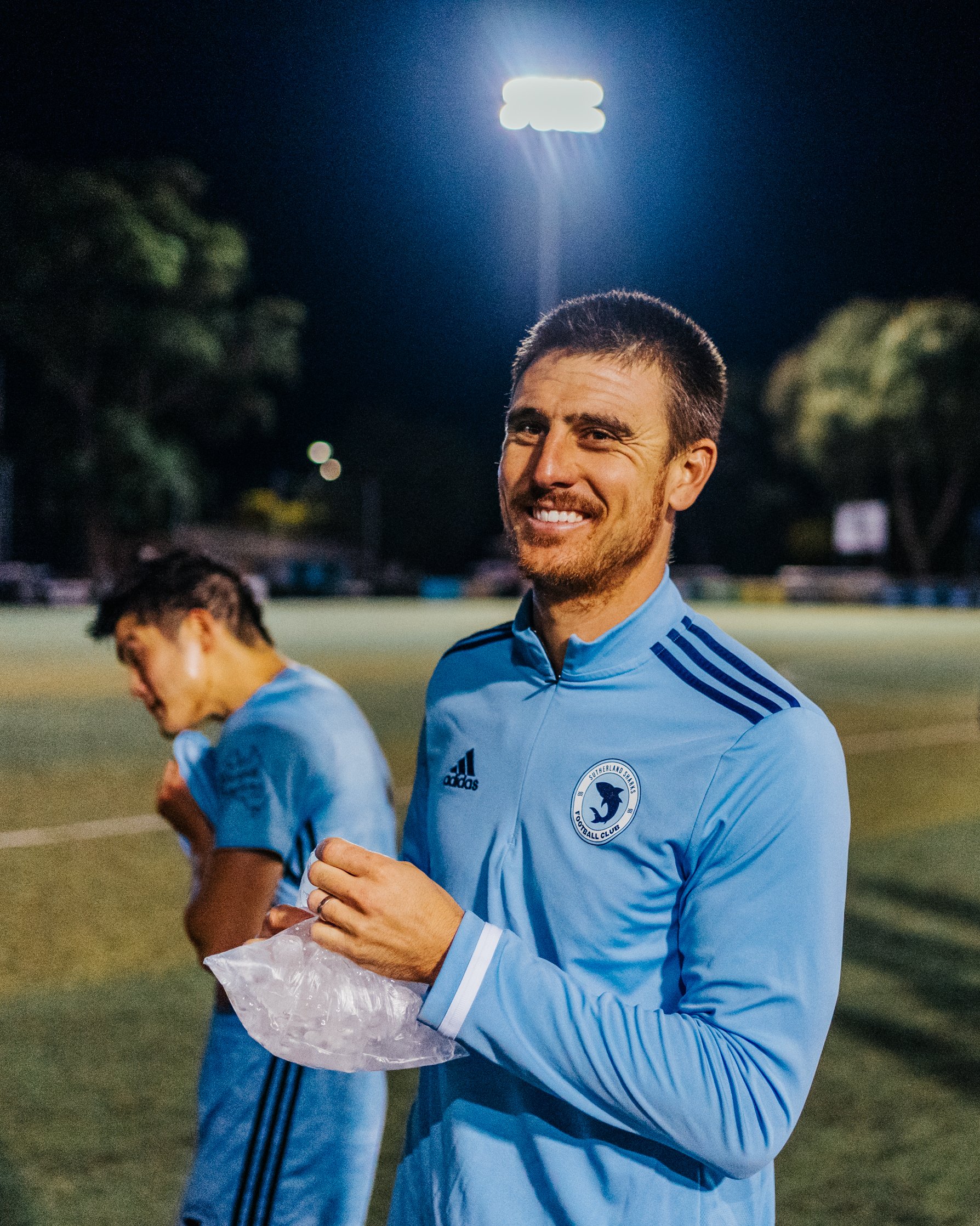 Two male soccer players in blue uniforms standing on a field at night, with one smiling at the camera and holding a plastic bag, the other looking down, illuminated by bright stadium lights.