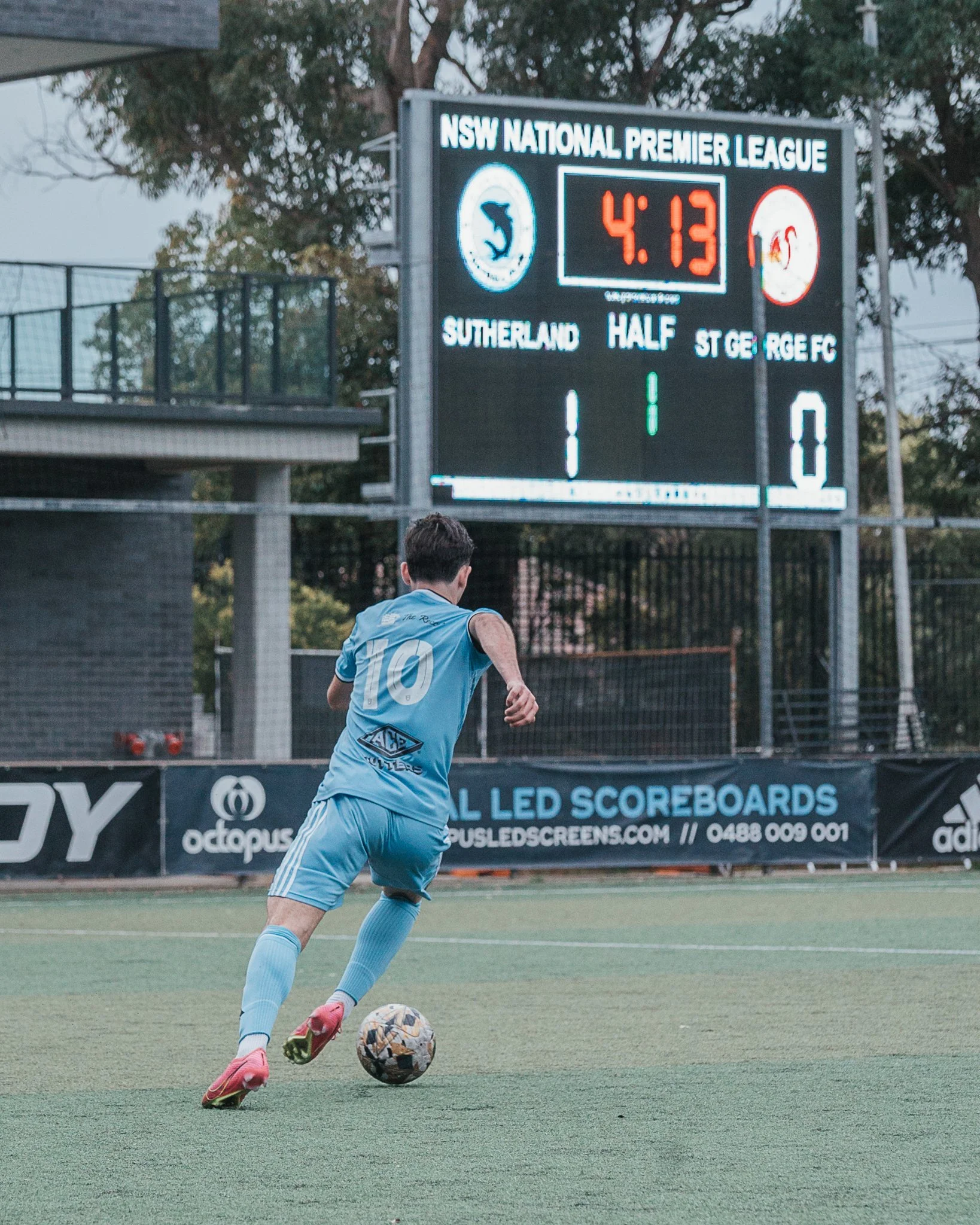 A young soccer player wearing a light blue uniform with the number 10 is running with a soccer ball during a match on a field. The scoreboard in the background shows Sutherland leading St George FC 1-0 at half-time, with 4 minutes and 13 seconds remaining.