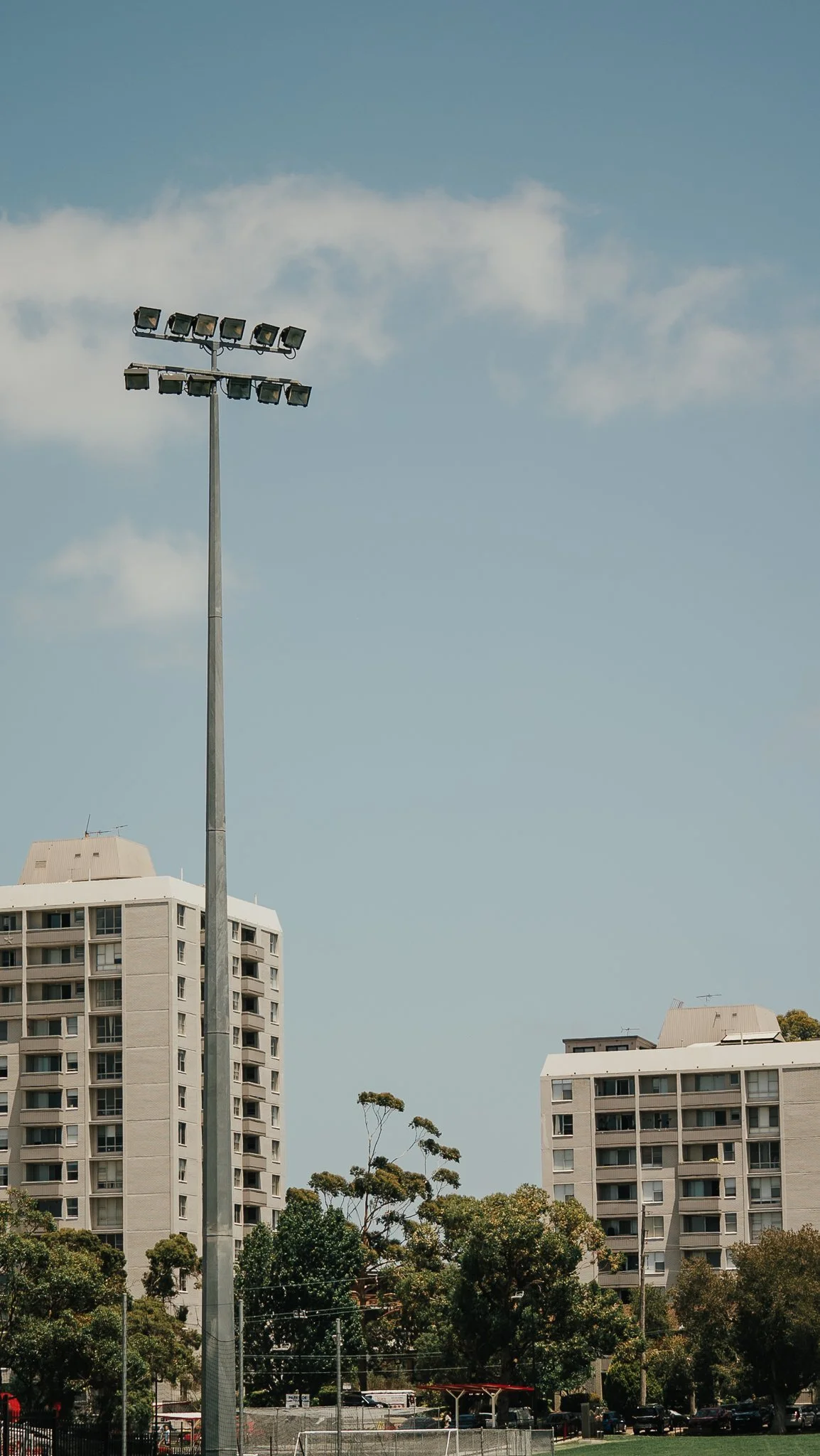 A tall outdoor stadium light pole with multiple floodlights, flanked by high-rise apartment buildings and trees under a partly cloudy sky.