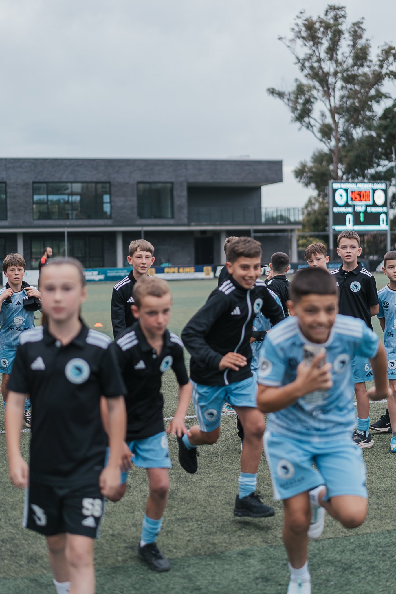 Children running on a soccer field during a game, with a scoreboard in the background.