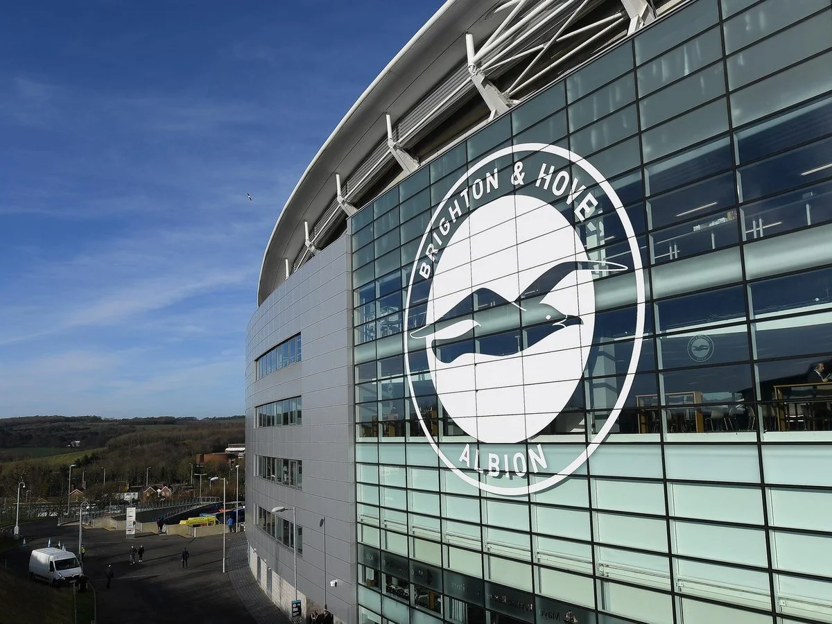 Exterior view of Brighton & Hove Albion football stadium with large glass facade and the club's logo featuring a seagull, under a blue sky.