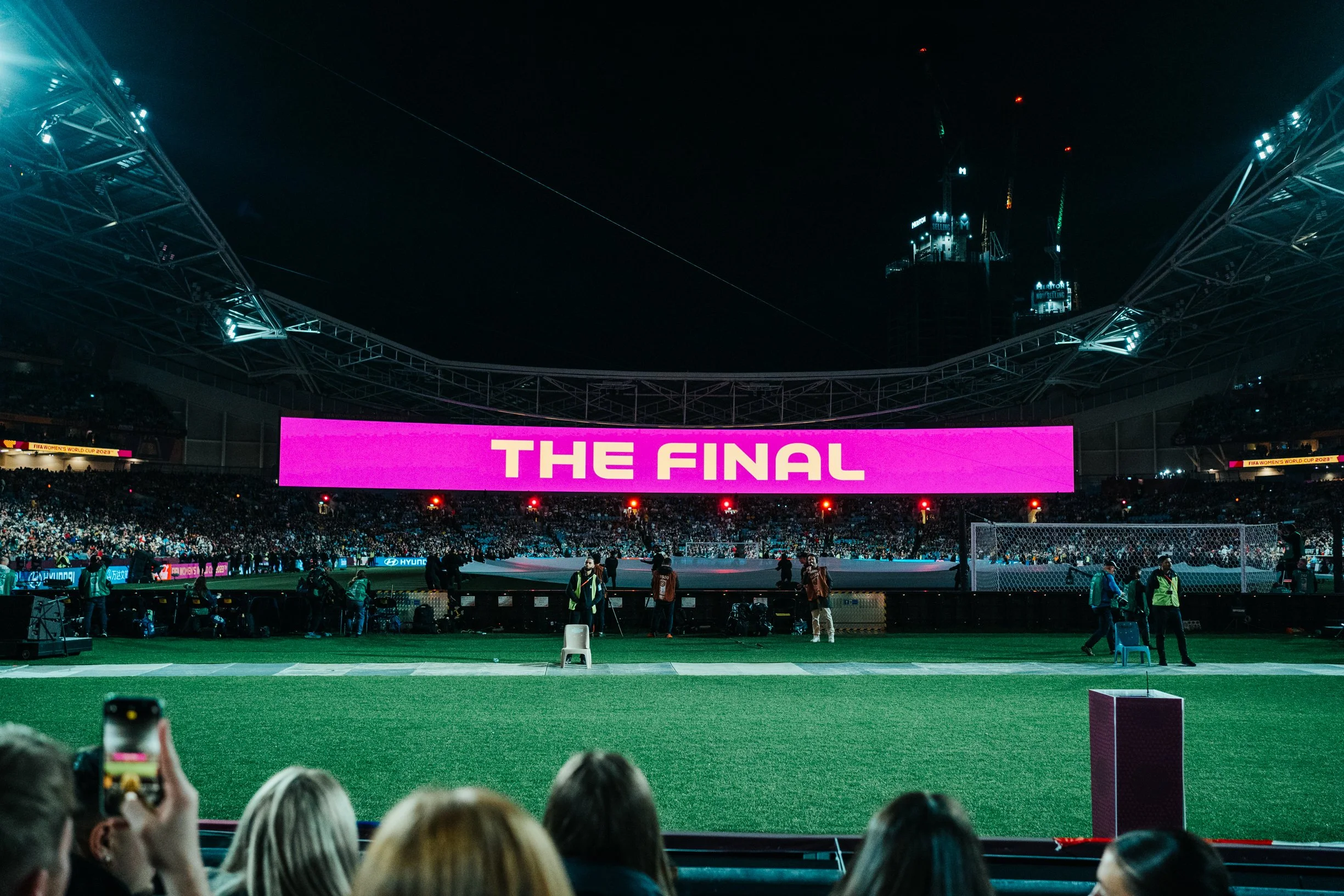 A stadium at night with a large pink LED screen displaying 'THE FINAL'. There are people on the field and in the stands, with some taking photos. Lights and a tall building can be seen in the background.