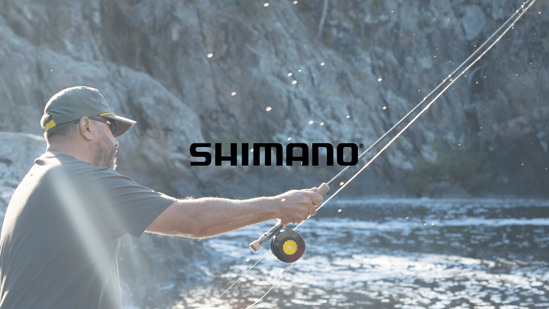 Man fishing in a river with rocky cliffs in the background, wearing sunglasses and a cap, holding a fishing rod