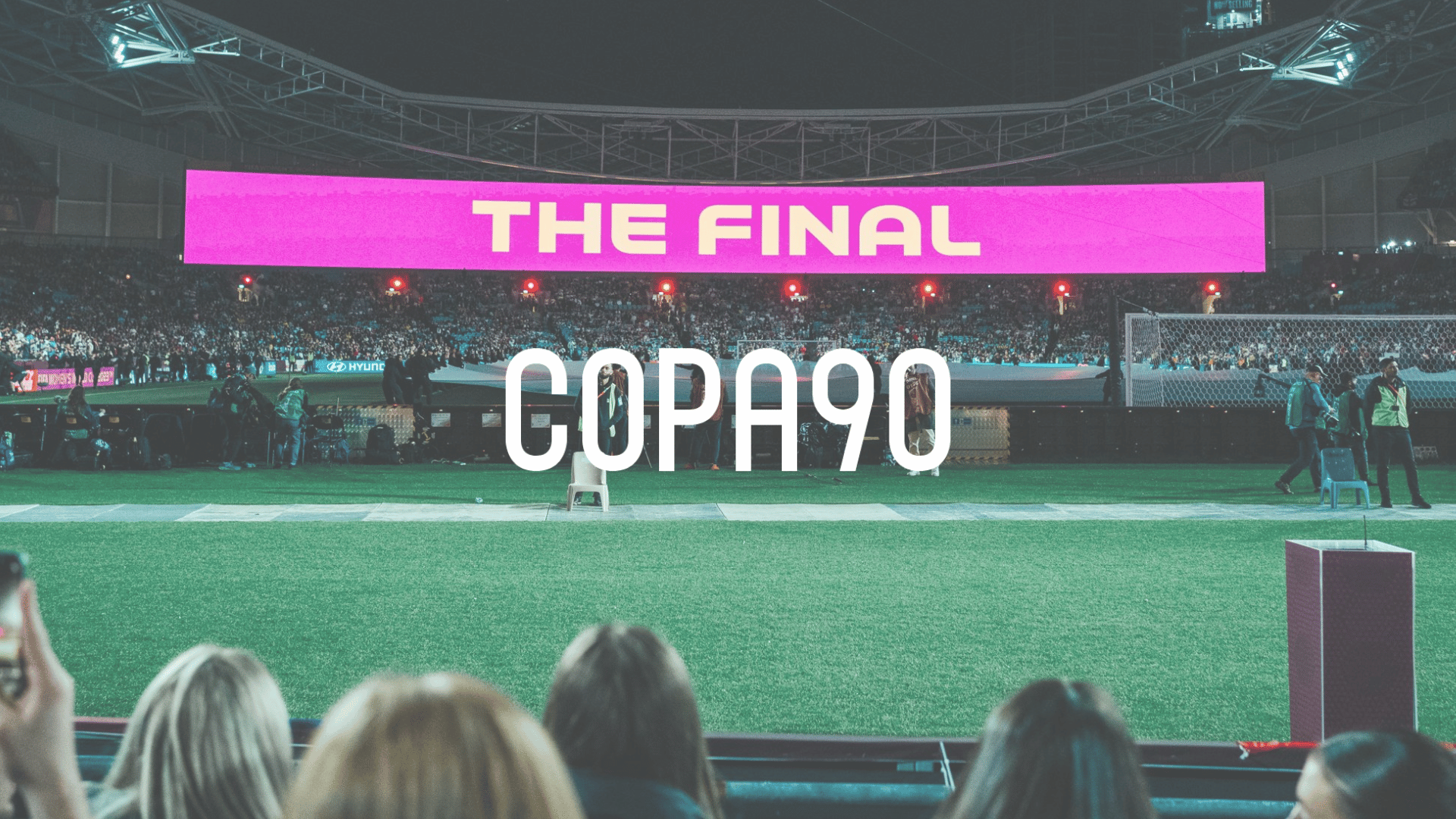 A stadium filled with spectators during nighttime, with a large electronic banner displaying 'THE FINAL' in pink and white, and another text overlay reading 'COPA90'.