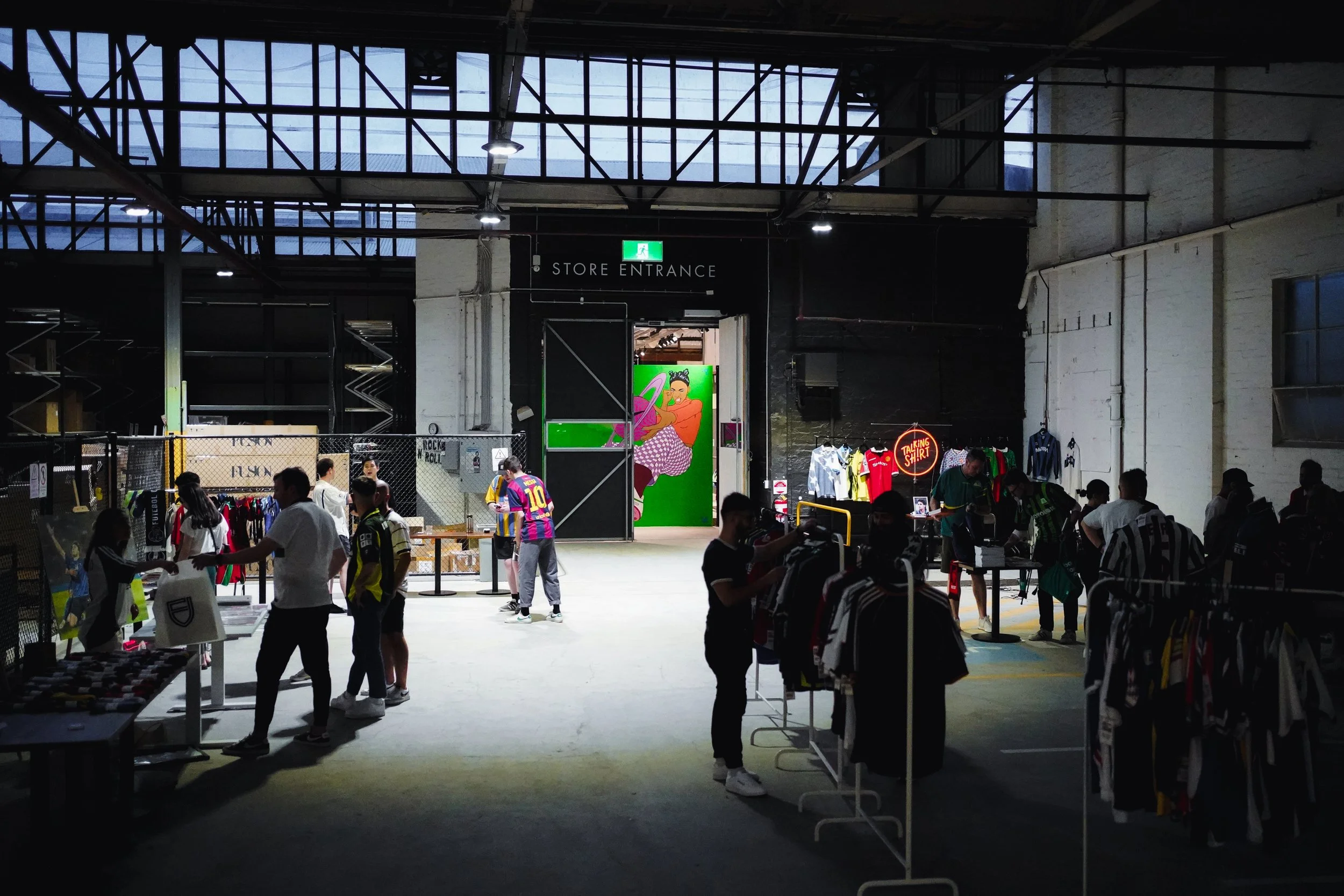 Inside a clothing store with racks of apparel, shoppers browsing, and colorful signage, including a green door with artwork and a neon sign that reads 'TALKING SHIRT.'