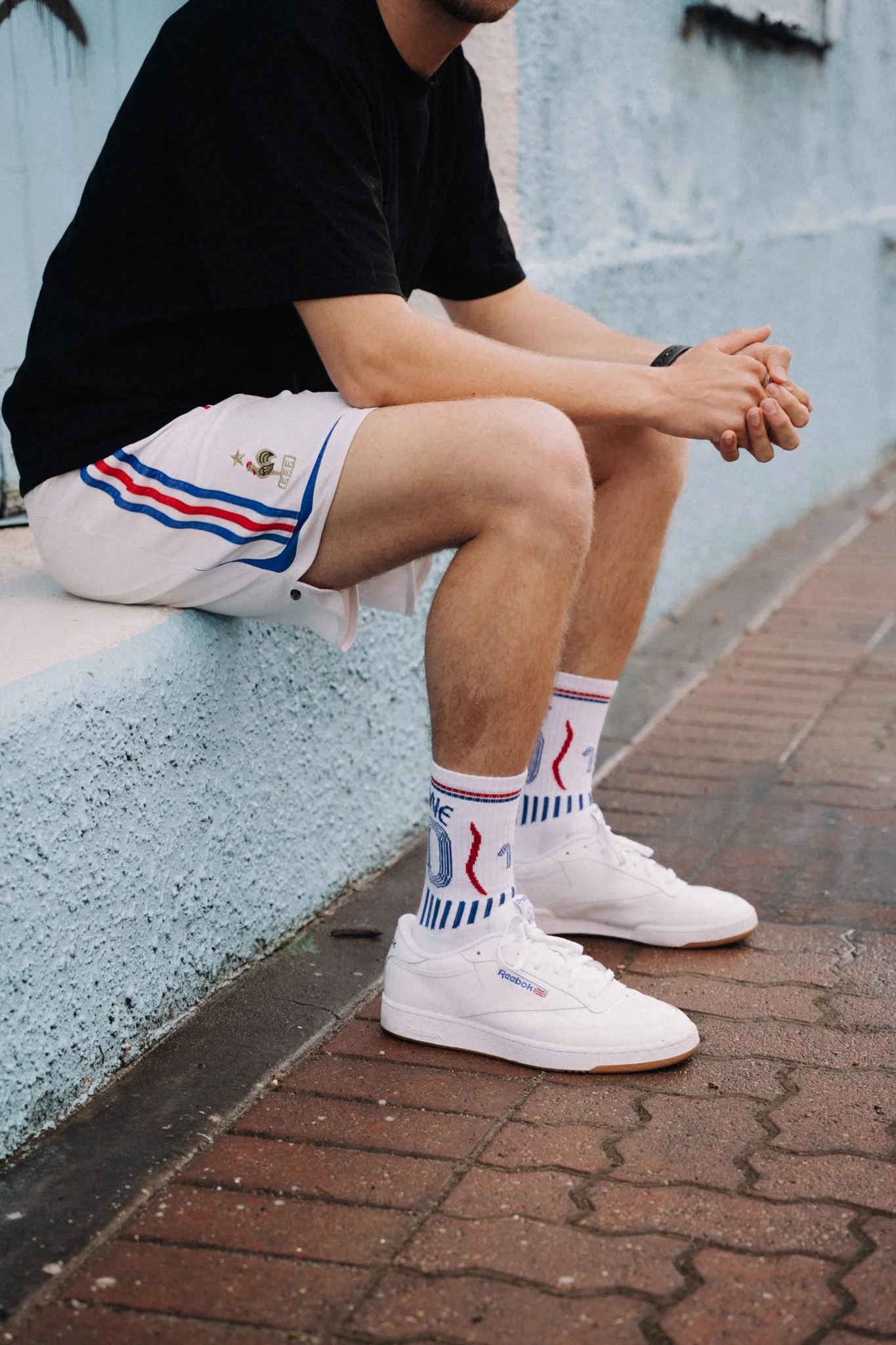 A person sitting on a concrete ledge, wearing a black t-shirt, white athletic shorts with blue and red stripes, white Reebok sneakers, and patterned athletic socks.