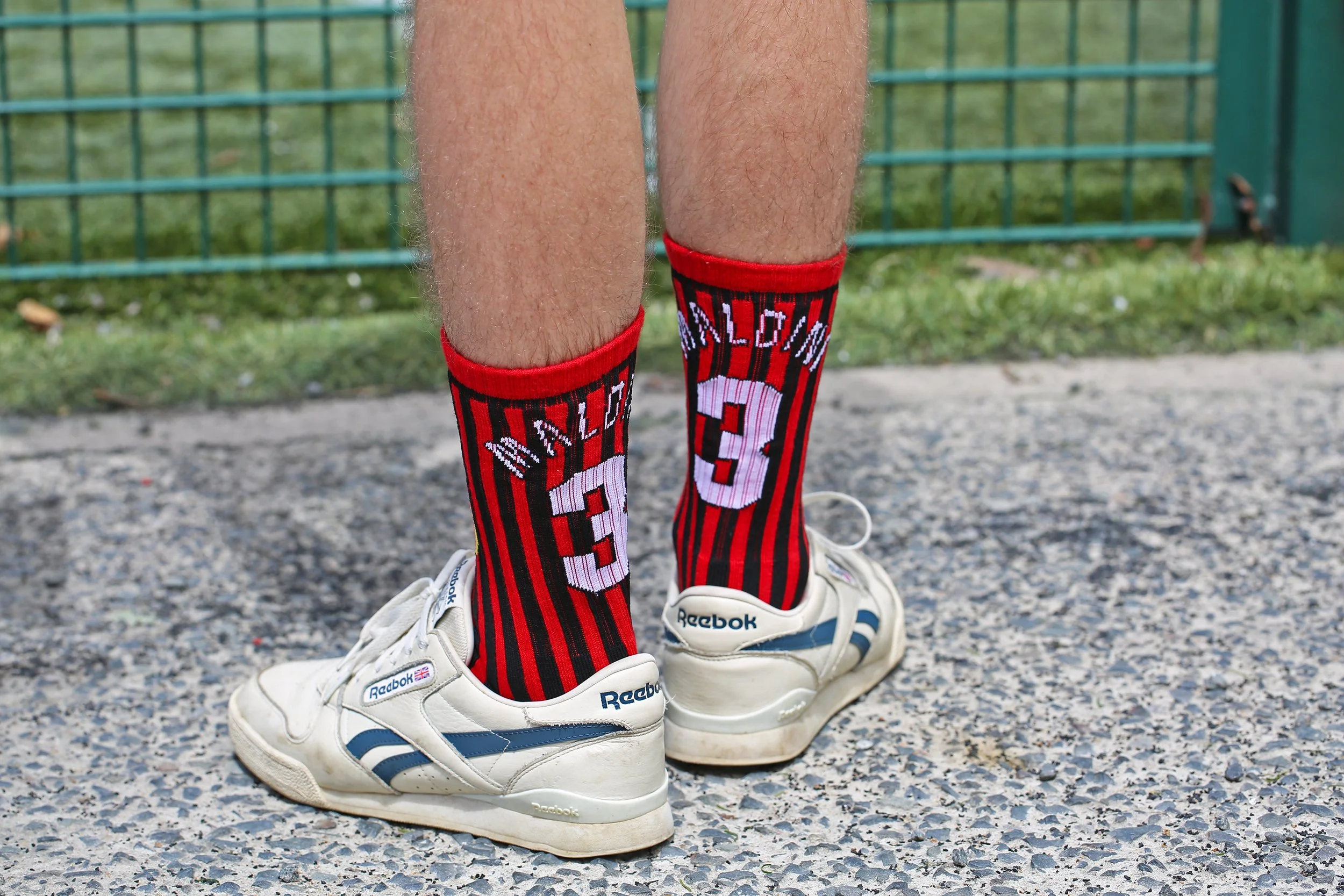 Person wearing white Reebok sneakers and red and black striped socks with the words "WILTON" and the number "3" on them, standing on a gravel surface near a green metal fence.