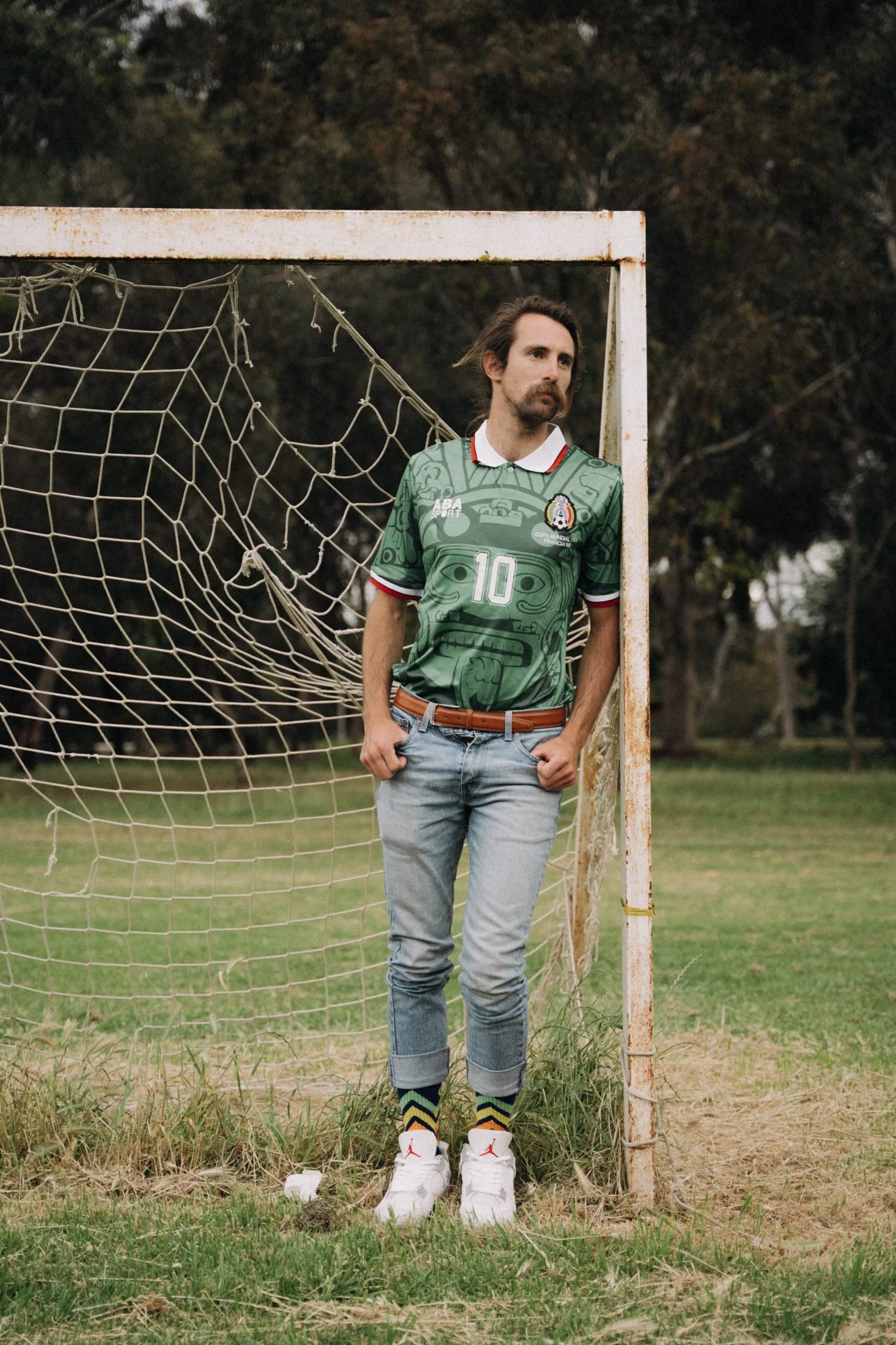 A man with long hair and a mustache in a green sports jersey with the number 10, standing in a rusty soccer goal on a grassy field with trees in the background.