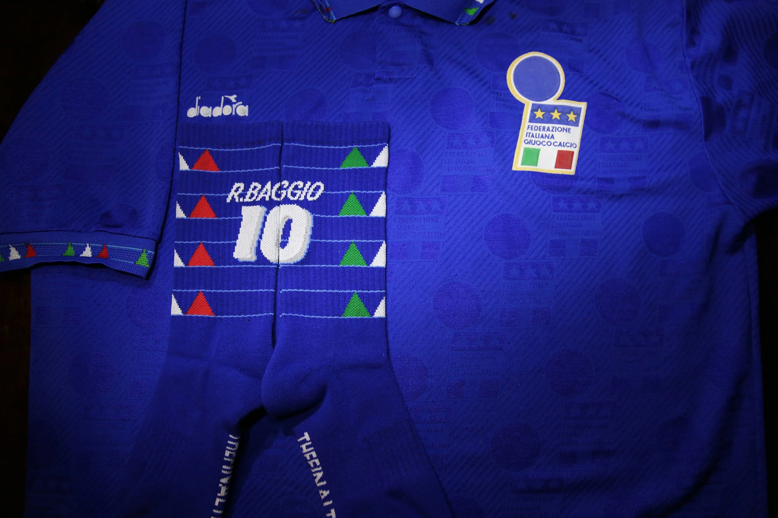 Italian soccer jersey with patches, a rolled pair of matching socks with colorful triangles, and a badge indicating Italian federation and goalkeeper team, all on a dark surface.