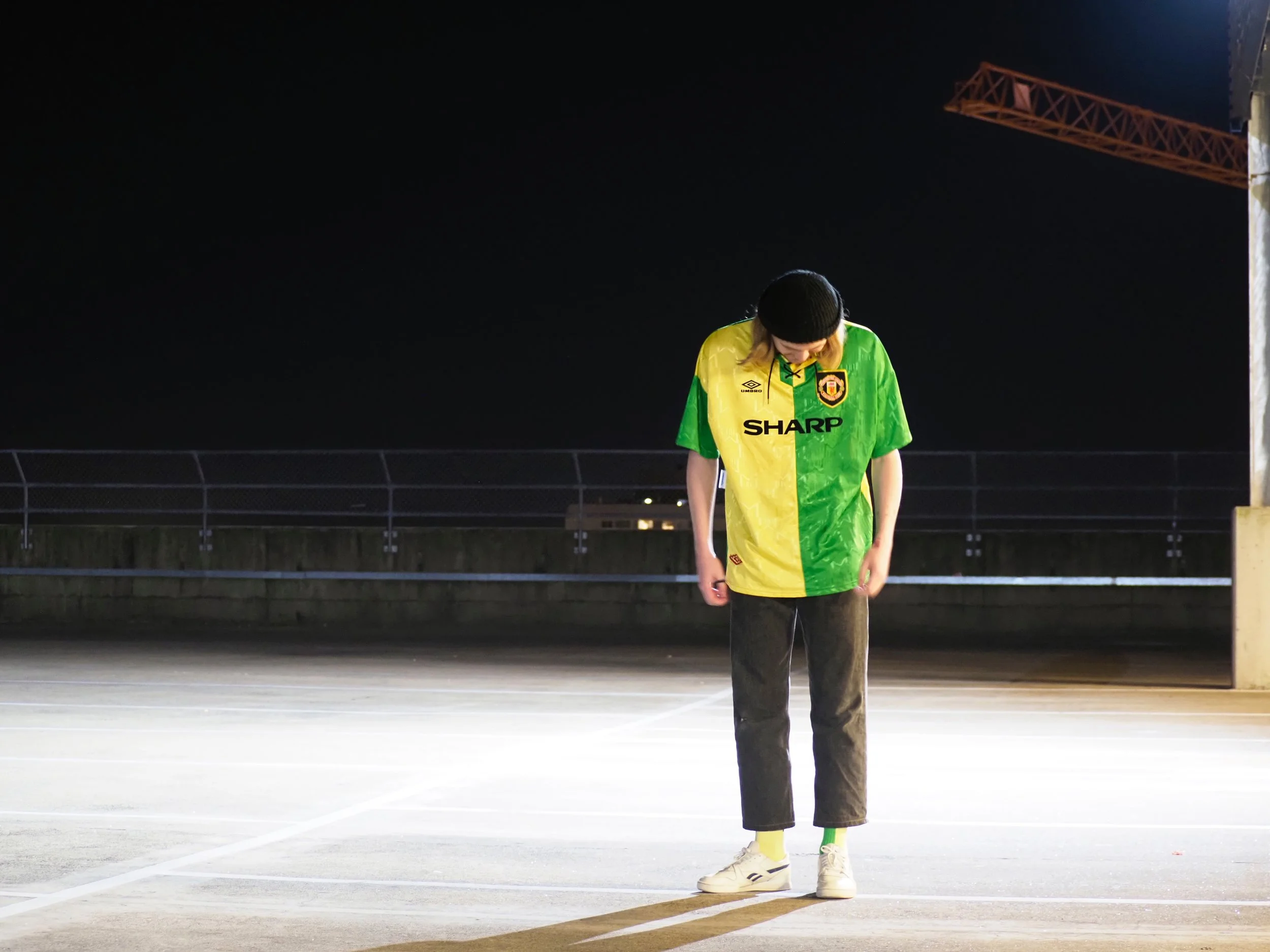 Person wearing a yellow and green sports jersey standing alone on an empty parking lot at night, looking down.
