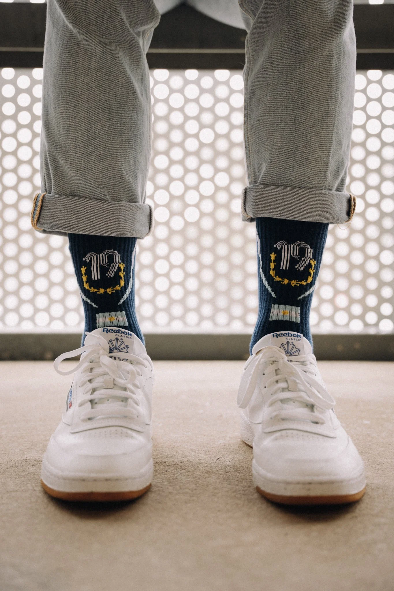 Person wearing rolled-up gray jeans, navy Reebok socks with beer mug design, and white Reebok sneakers, standing on beige carpet with a perforated metal background.