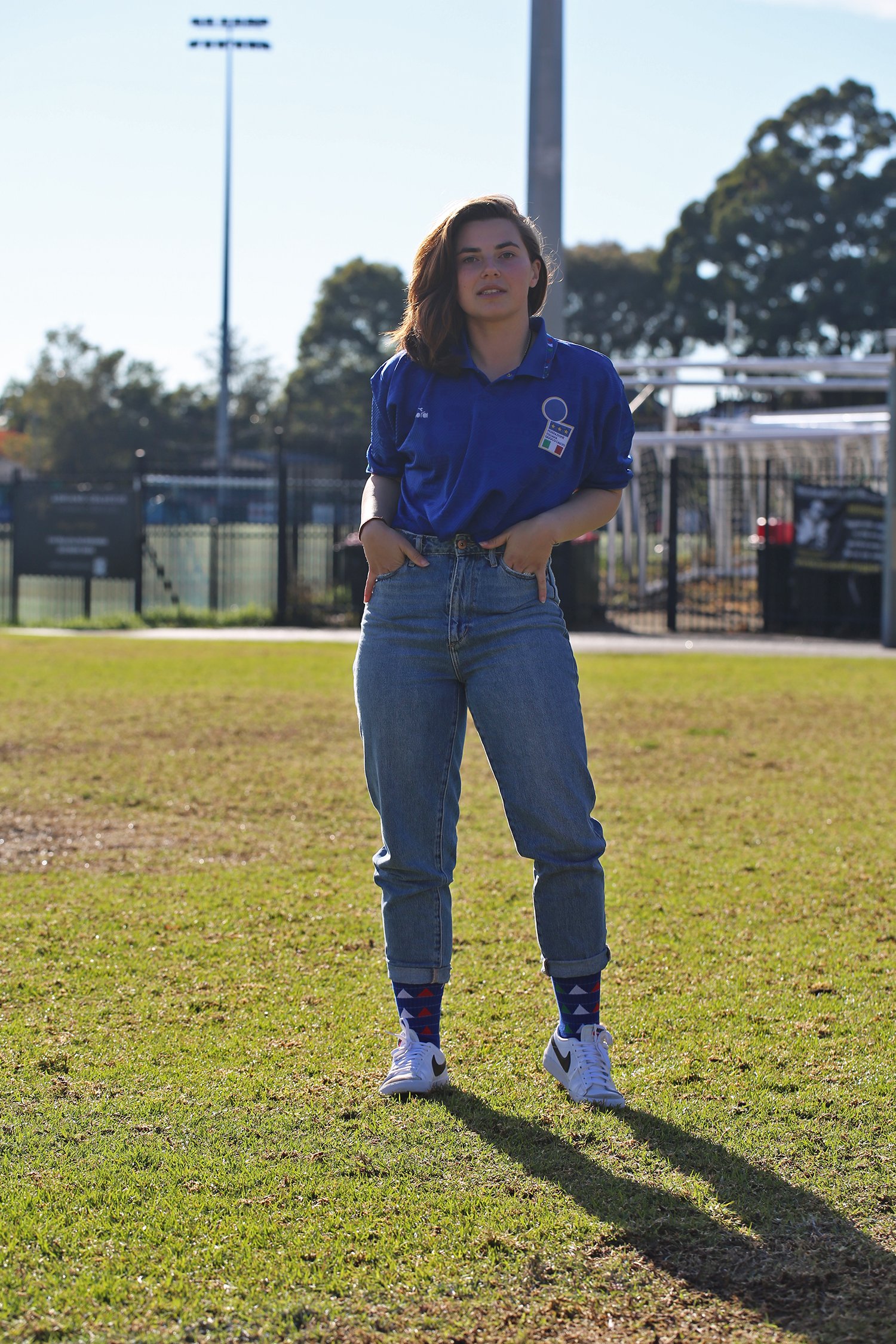A young woman with short dark hair standing on grass field in a sports stadium, wearing a blue sports jersey with a logo, high-waisted jeans, white sneakers, and colorful socks, with a sports field and trees in the background.
