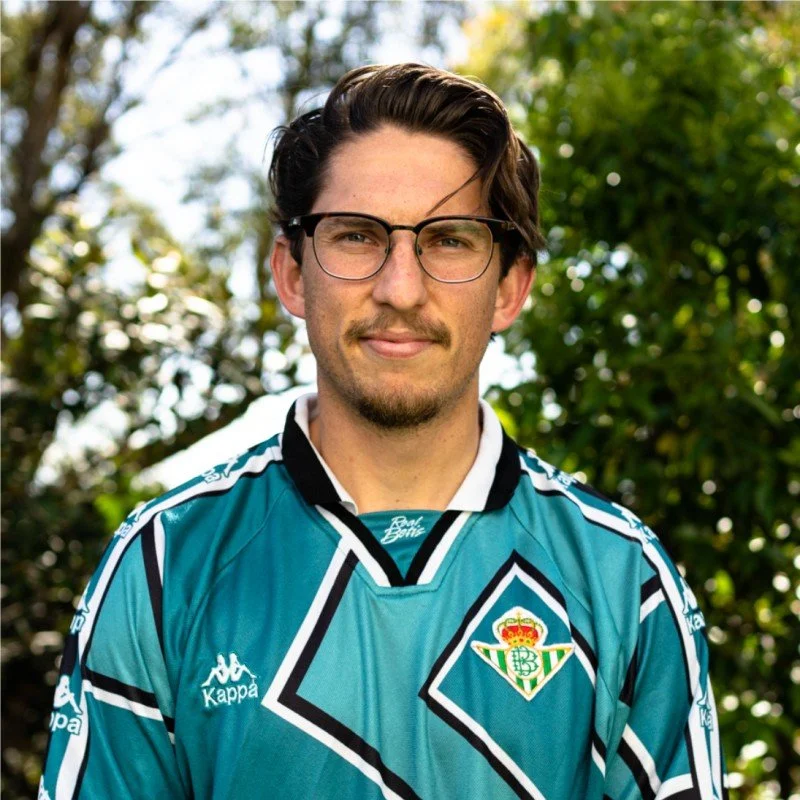 Young man wearing glasses and a sports jersey outdoors with green trees in the background.