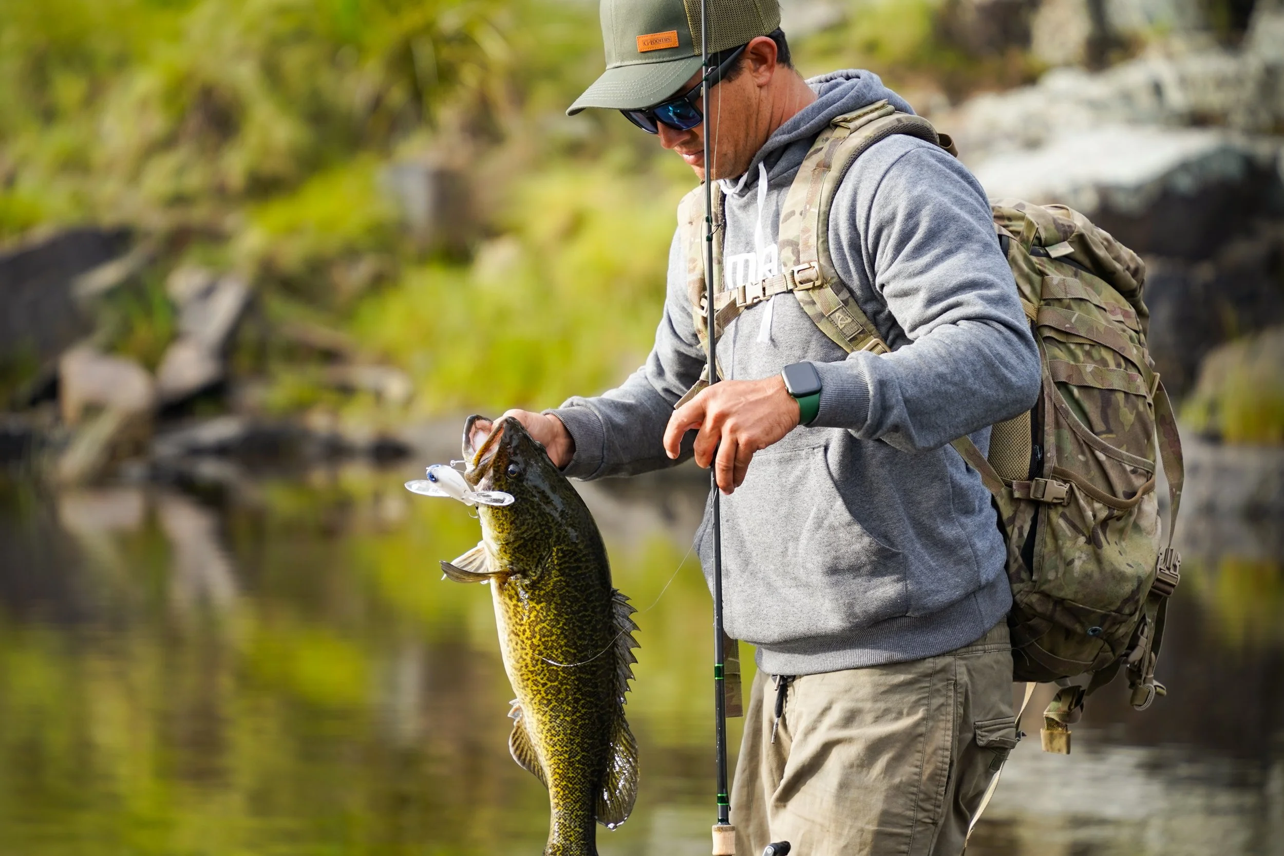 Man fishing in a river holding a large fish with a fishing lure in its mouth.