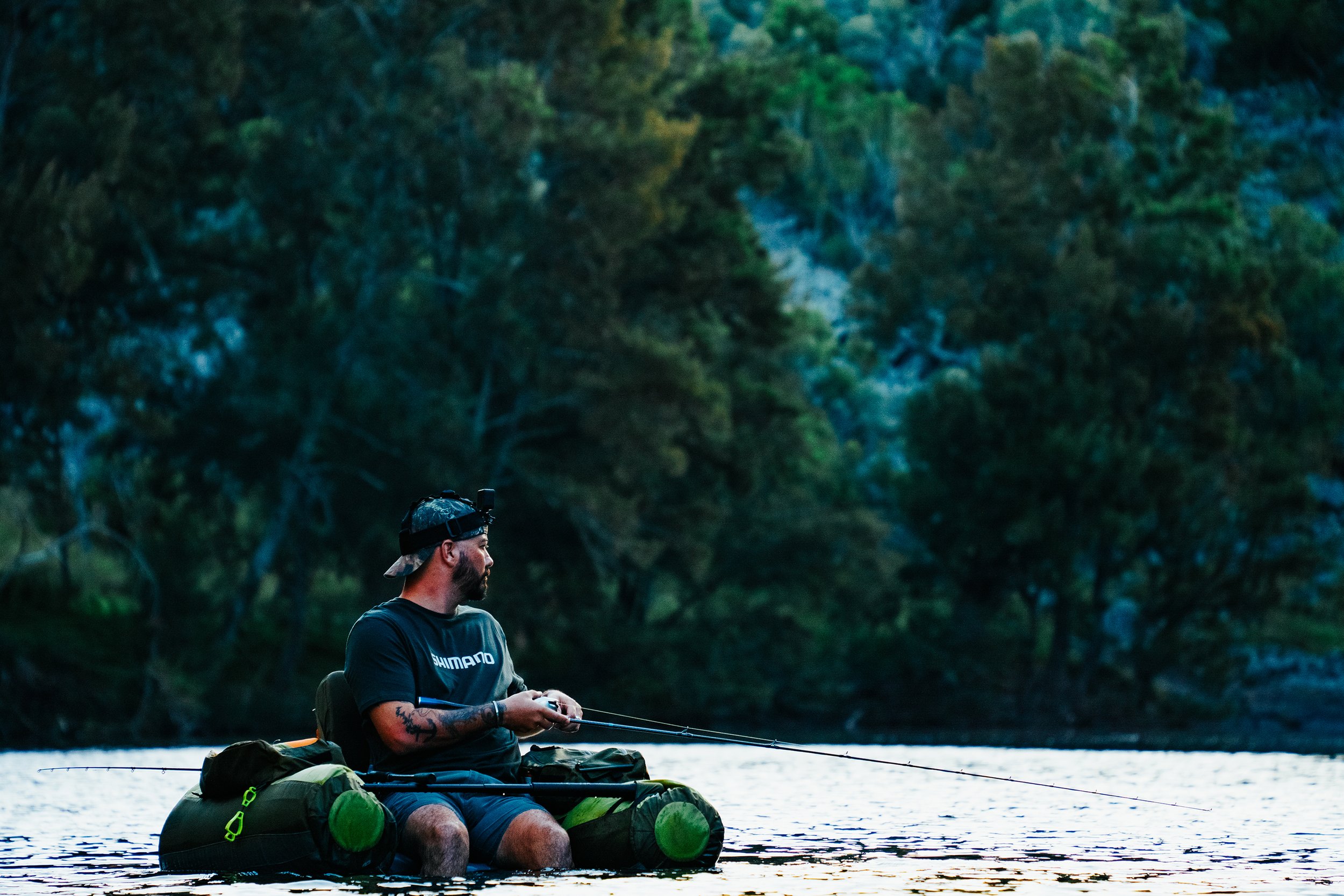 A man fishing in a lake during dusk, sitting on a kayak with gear, surrounded by trees.