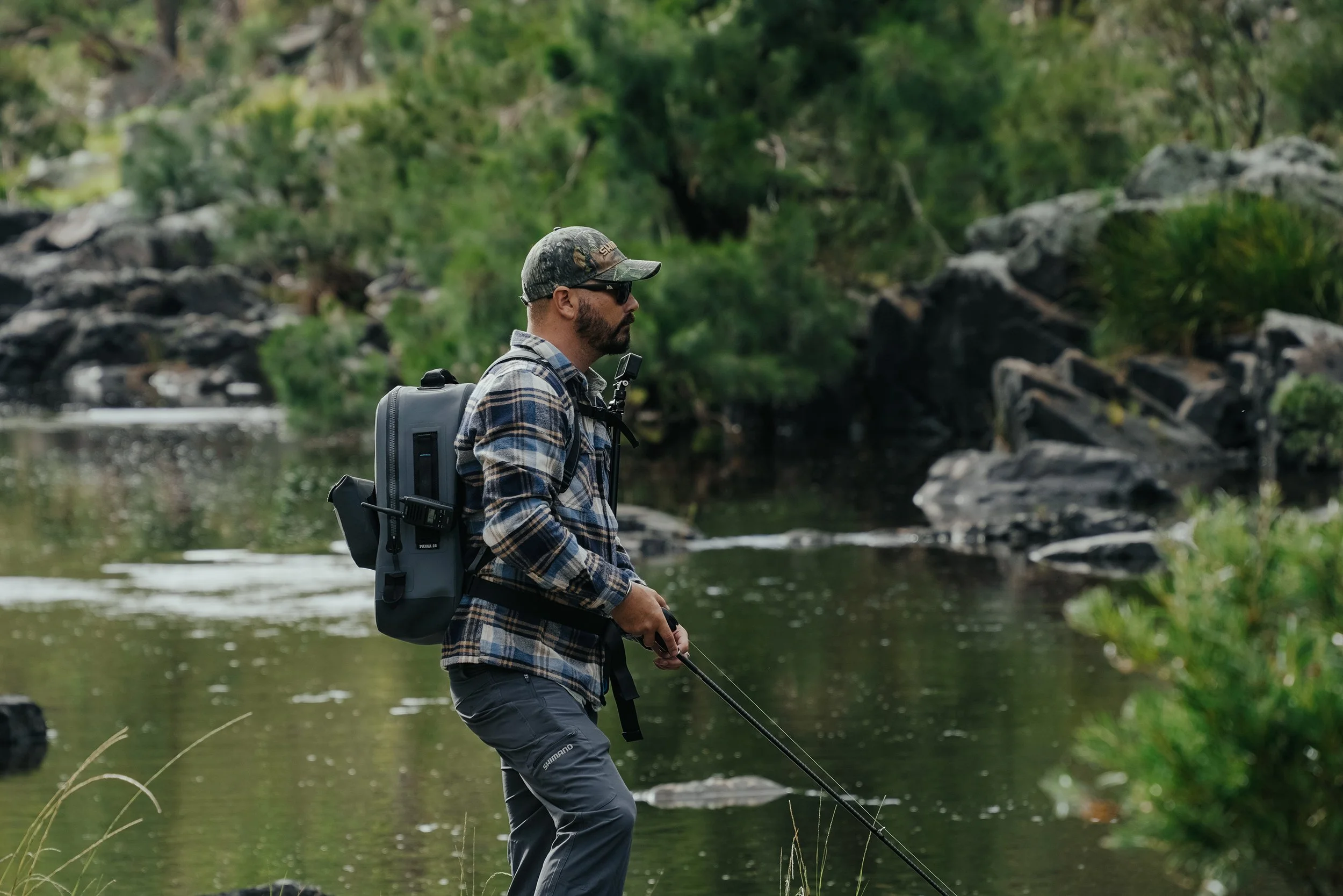 A man in outdoor gear stands on the edge of a creek, holding a fishing rod, surrounded by green trees and rocks.