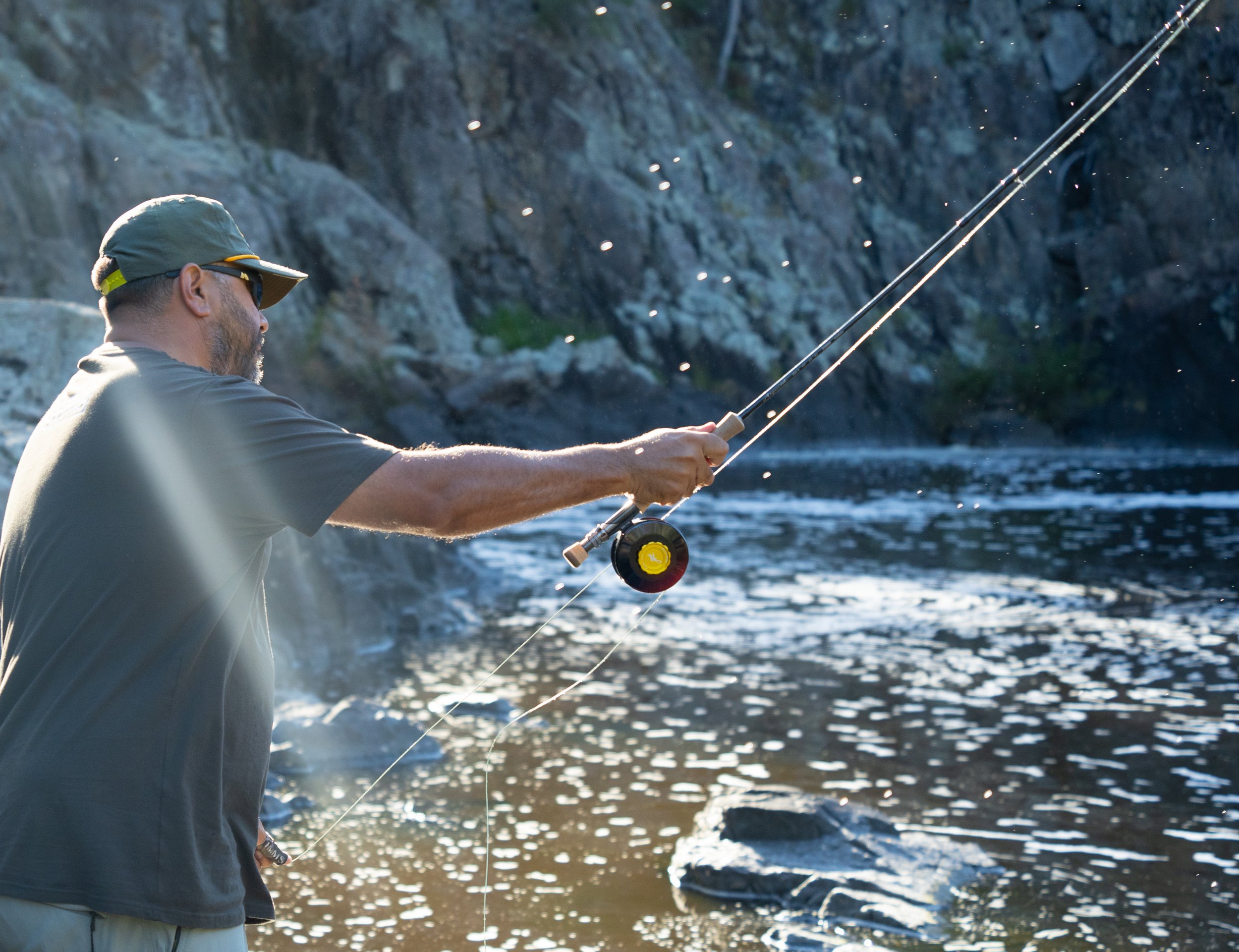 A man fishing in a river with rocky cliffs in the background, wearing a gray cap and T-shirt, holding a fishing rod.