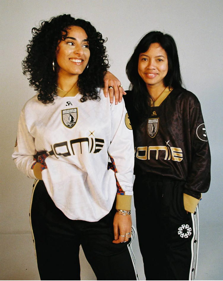 Two women standing next to each other, wearing sports jerseys with logos and black pants with white stripes, smiling at the camera.