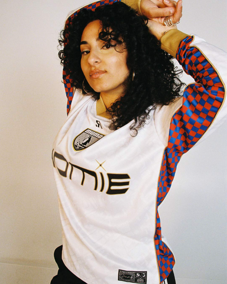 A woman with curly black hair posing with her right arm raised, wearing a white sports jersey with black and gold details and a patterned red and blue long sleeve shirt underneath.