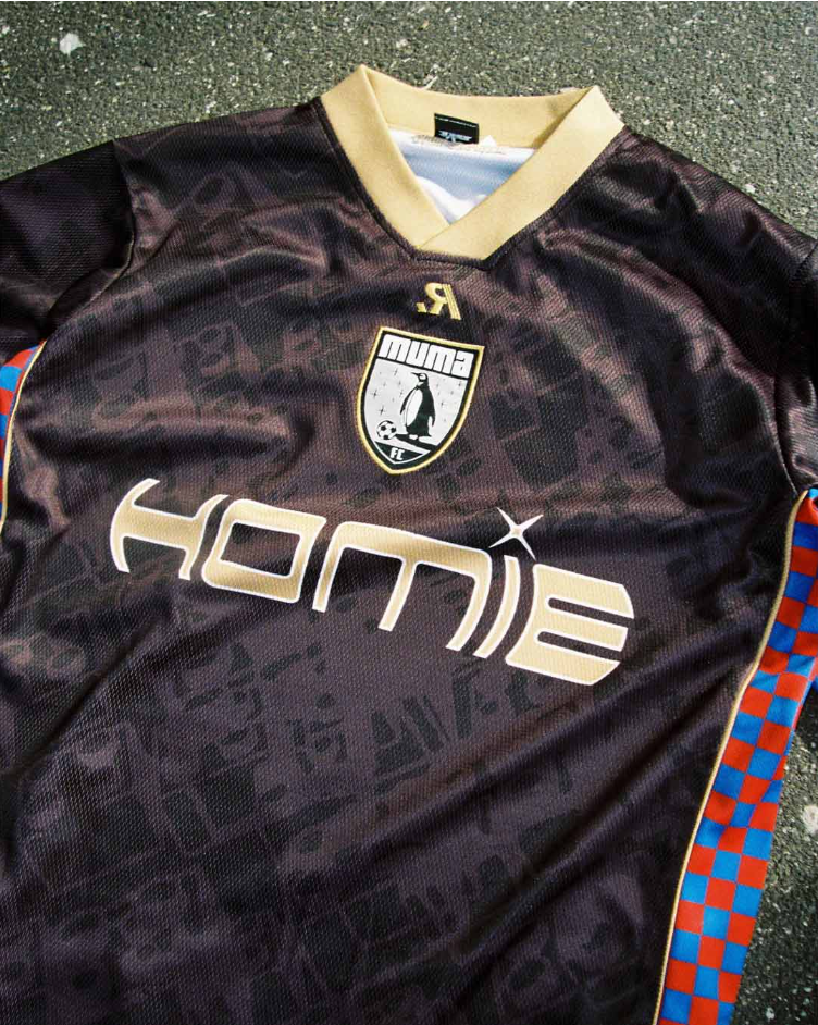 A dark-colored soccer jersey with the Croatian checkered pattern on the sleeves, a gold collar, and a logo featuring a penguin playing soccer with the word "Puma" above it, and the word "HOMIE" displayed prominently across the front.
