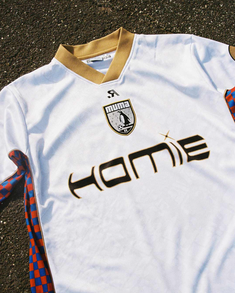 A white football jersey with gold collar details laid out on dark pavement. It features a PUMA logo, a badge with a penguin and the text 'numa FC,' and the word 'homie' across the front in black lettering.