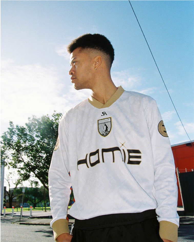 A young man with short dark hair wearing a white football jersey with gold accents, standing outdoors against a blue sky and trees.