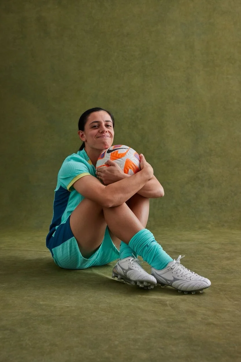 Female soccer player sitting on the ground, hugging a soccer ball, wearing teal athletic uniform and white cleats, against a plain green backdrop.