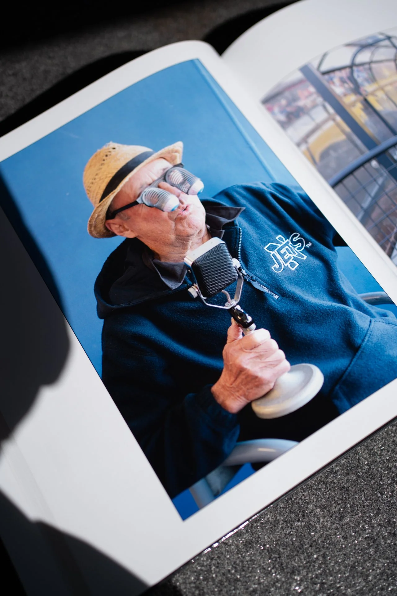 A man wearing a hat and a hoodie with a logo, holding a microphone, and sporting binoculars over his eyes, sitting on a chair against a blue background.