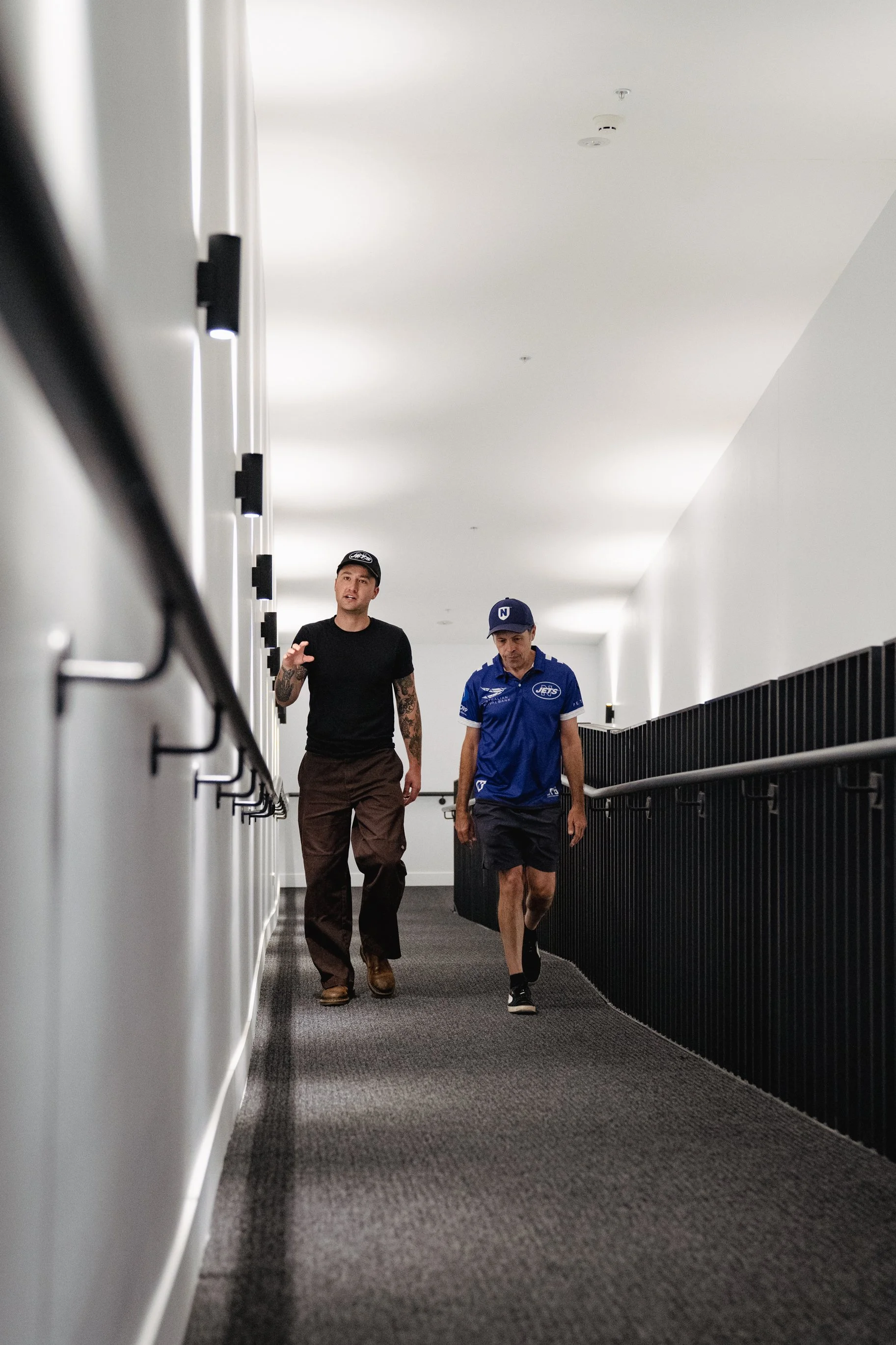Two men walking down a hallway with black railings and white walls, one wearing a black shirt and brown pants, the other in a blue sports shirt and black shorts.