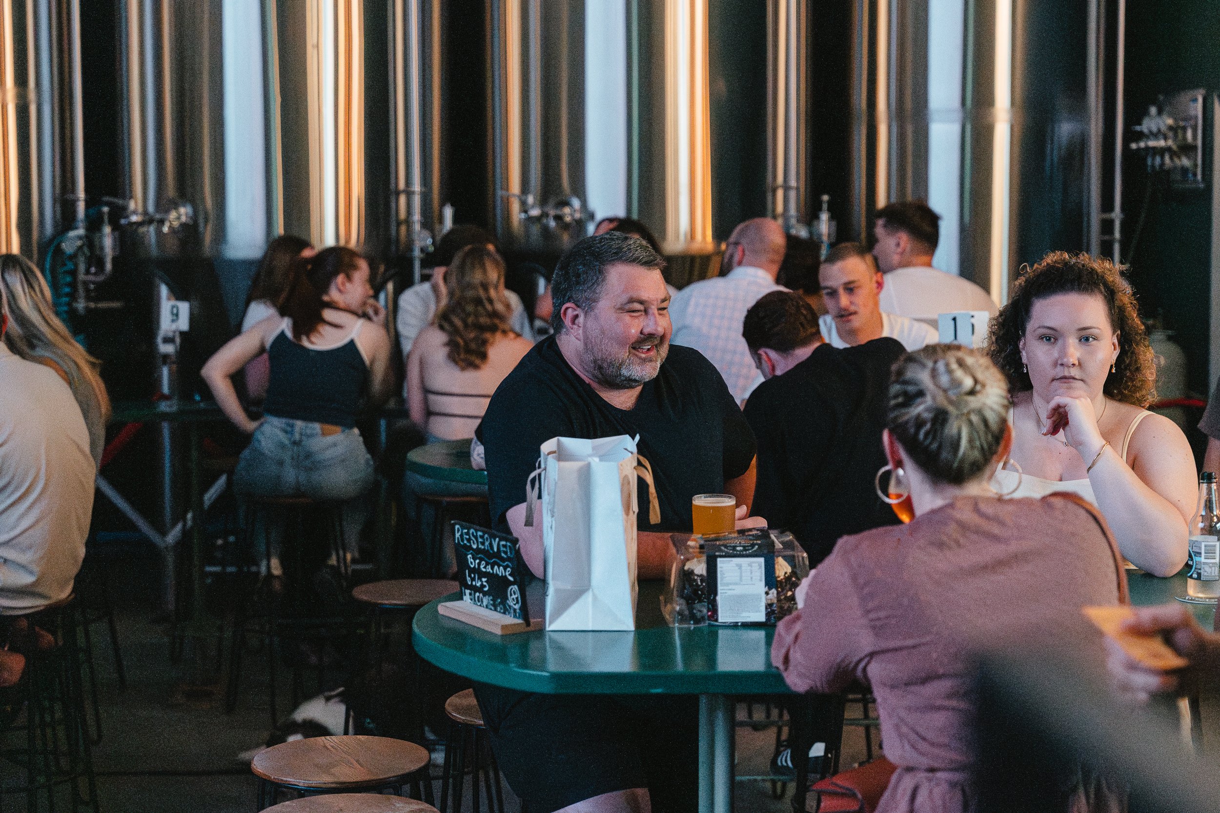 People socializing at a bar or brewery, with large metal tanks in the background and a reserved sign on the table.