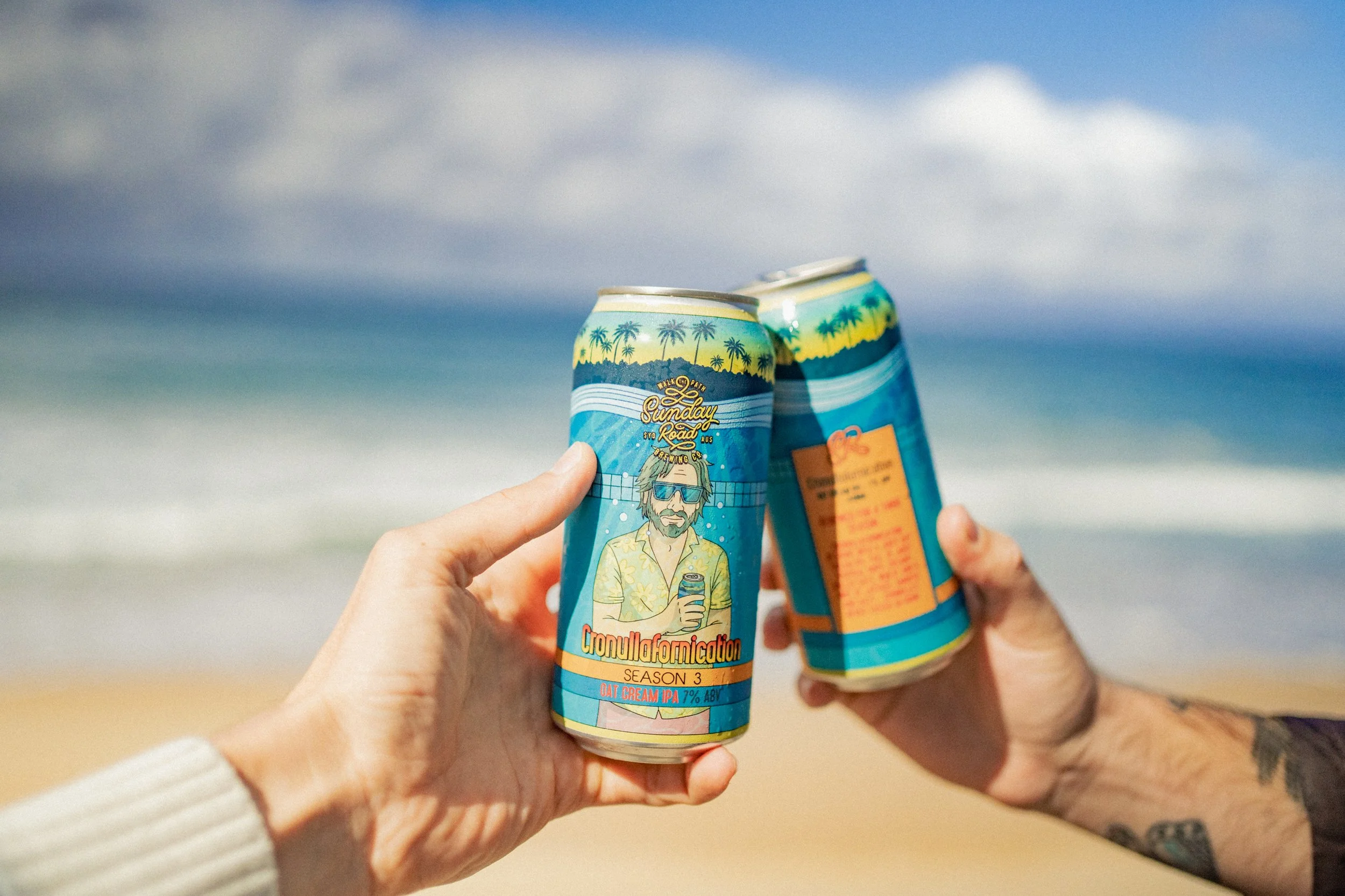 Two hands holding cans of Cronulla forniccation season 3 oat cream IPA at the beach with ocean and cloudy sky in the background.