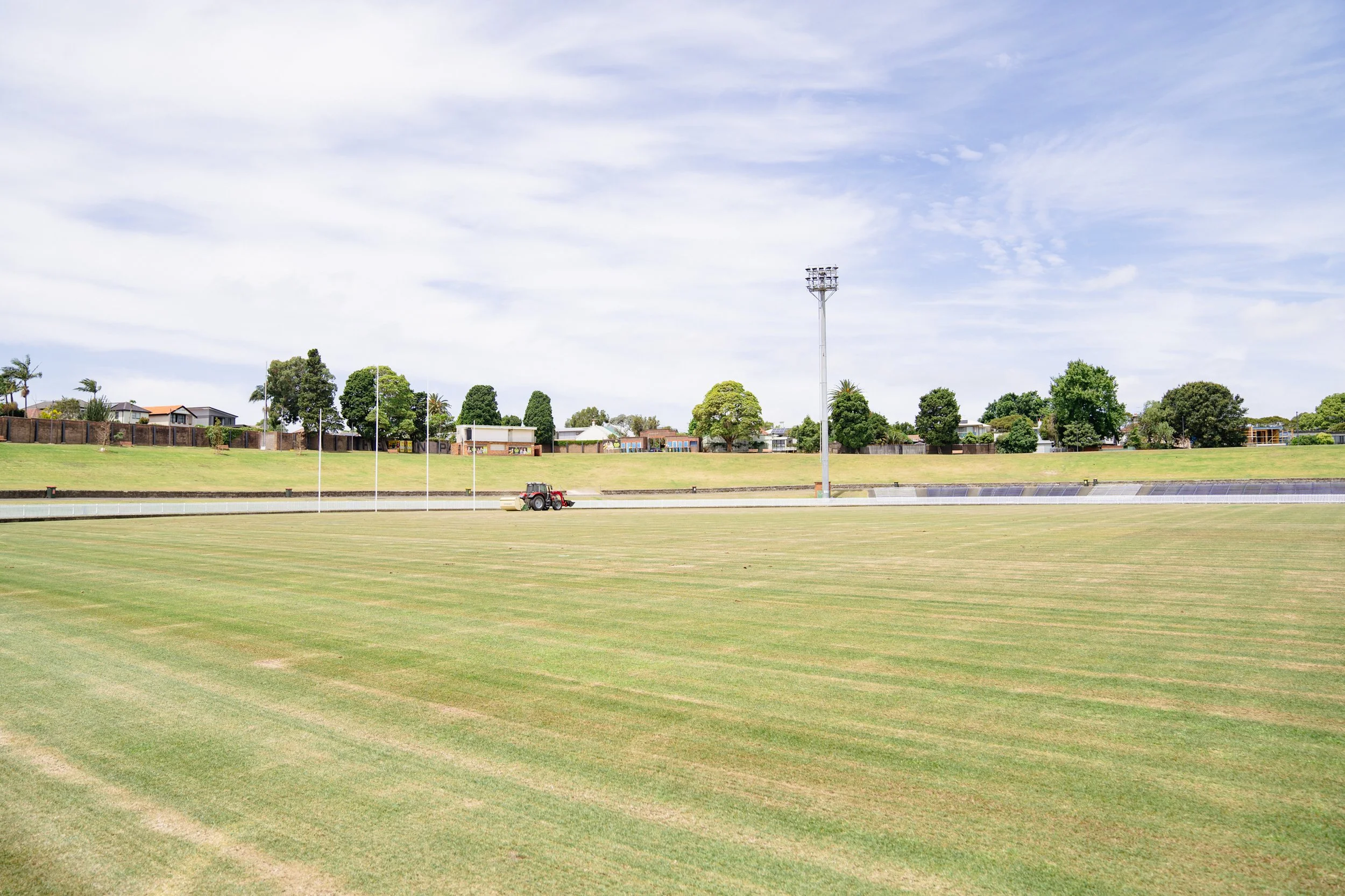 An empty outdoor sports field with freshly mowed green grass, goal posts, and a tractor nearby. Residential houses and houses with trees are visible in the background under a partly cloudy sky.