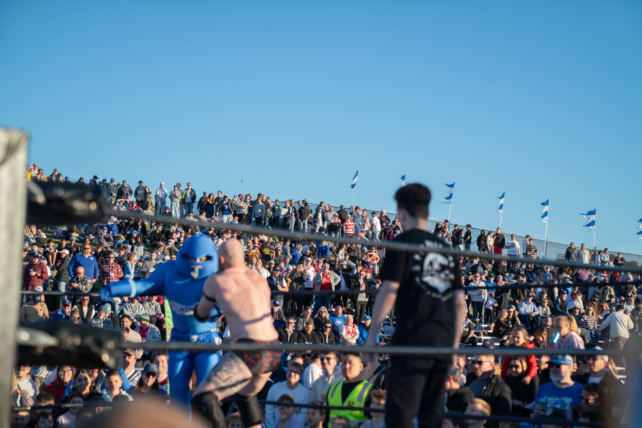 A wide view of a large crowd watching an outdoor boxing match taking place in a ring, with audience seated and standing on hills, some waving blue flags, under a clear blue sky.