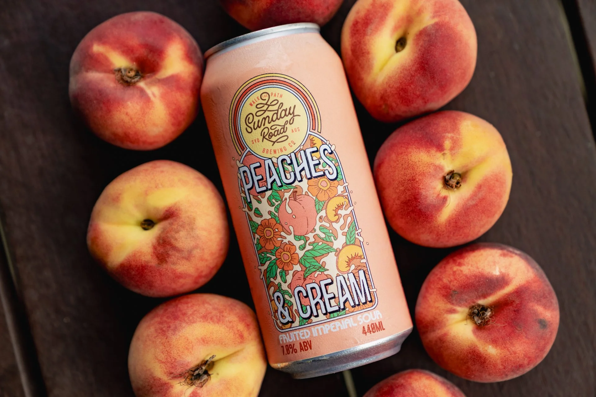 A can of Sunday Road Peaches & Cream flavored beer surrounded by seven peaches on a dark surface.