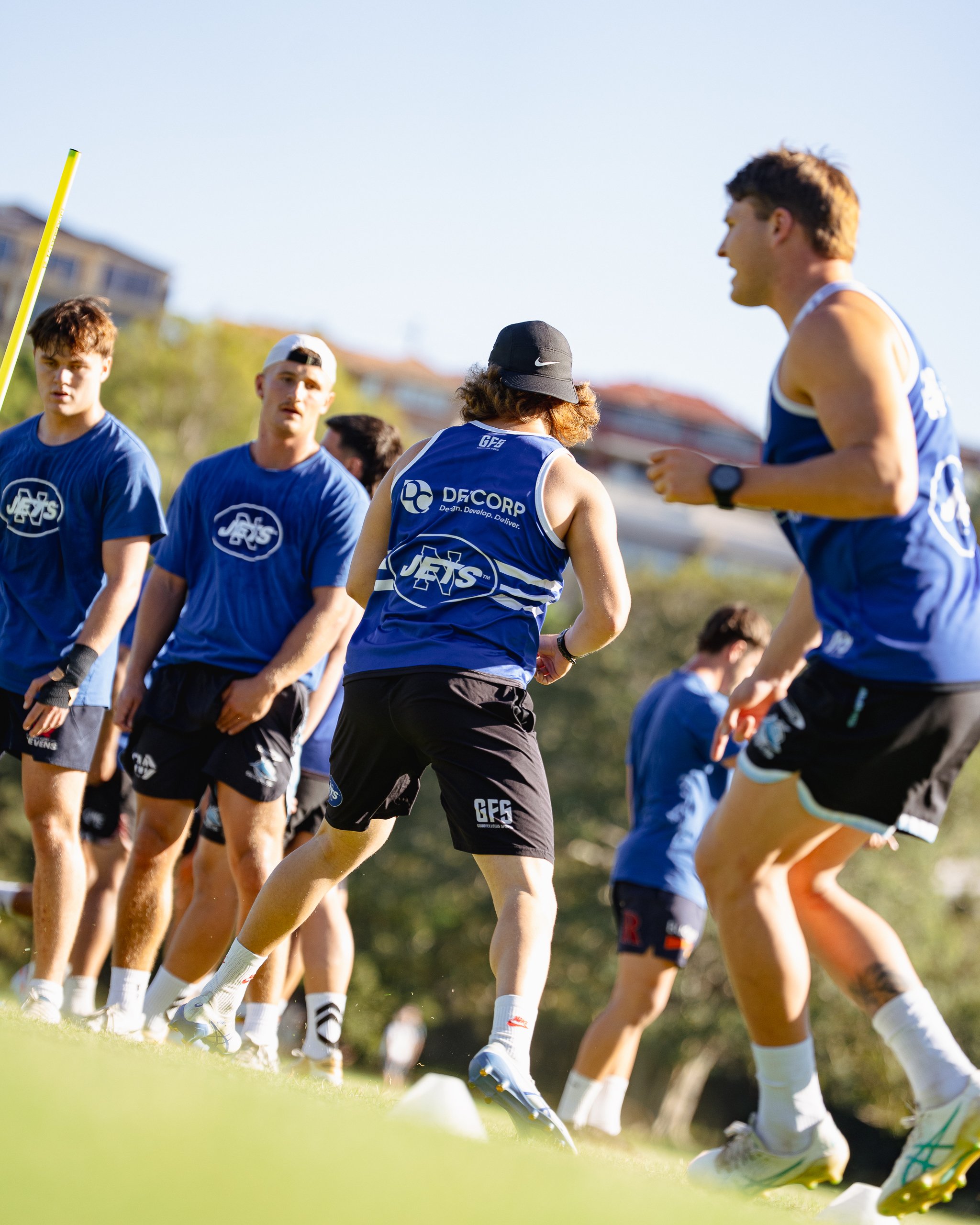Group of young athletes running during a training session outdoors on a sunny day, wearing blue sports jerseys.