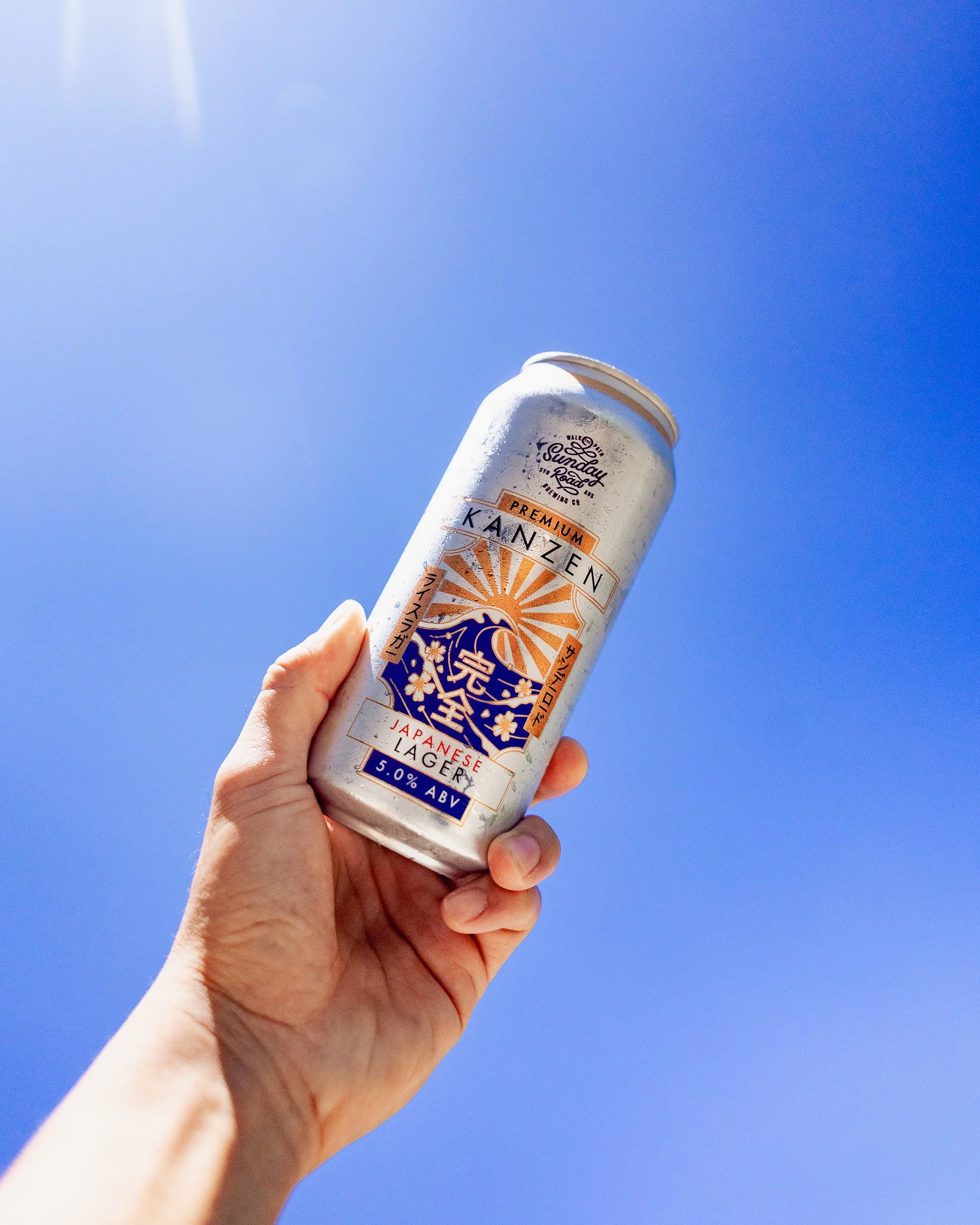 A hand holding a can of Japanese lager beer against a clear blue sky.