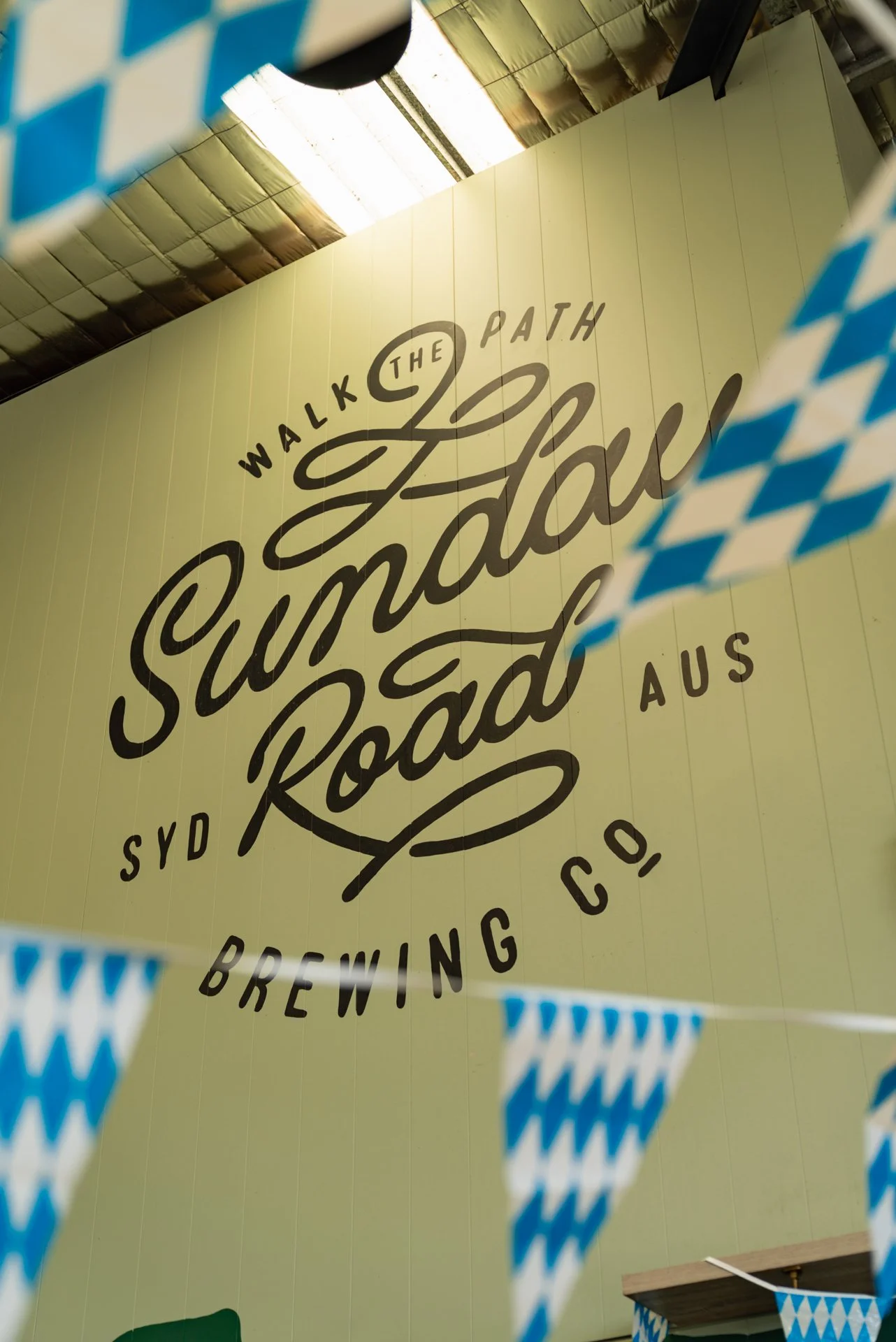 A large wall with the text 'Walk the Sunad Road' and 'Brewings Co' displayed prominently, partially obscured by blue and white checkered pennant banners hanging in the foreground.
