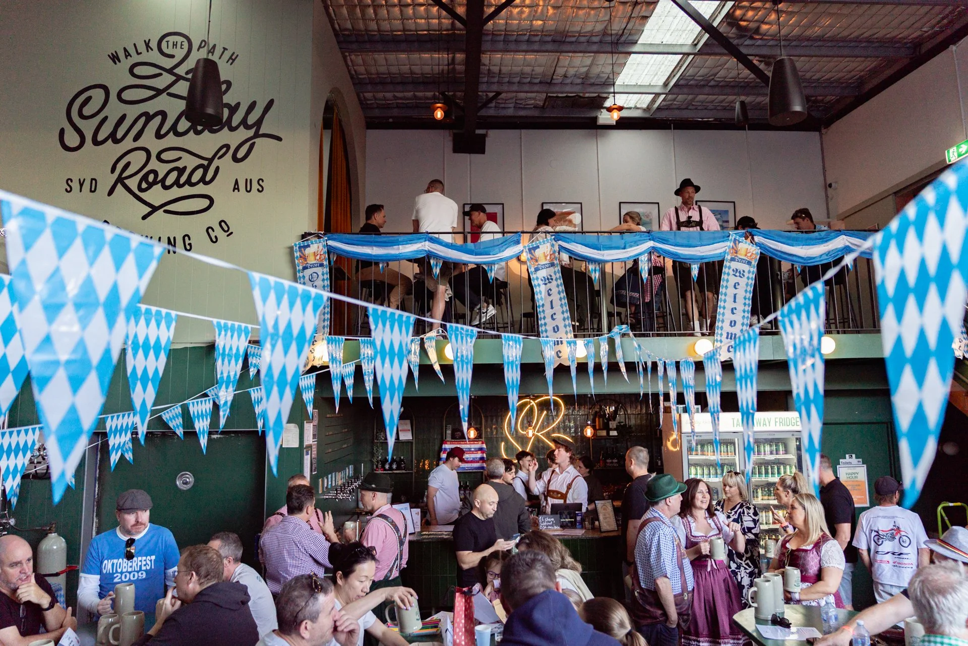 A crowded room decorated with blue and white checkered flags, celebrating Oktoberfest, with people dressed in traditional Bavarian clothing, socializing, and enjoying drinks at a festive event.