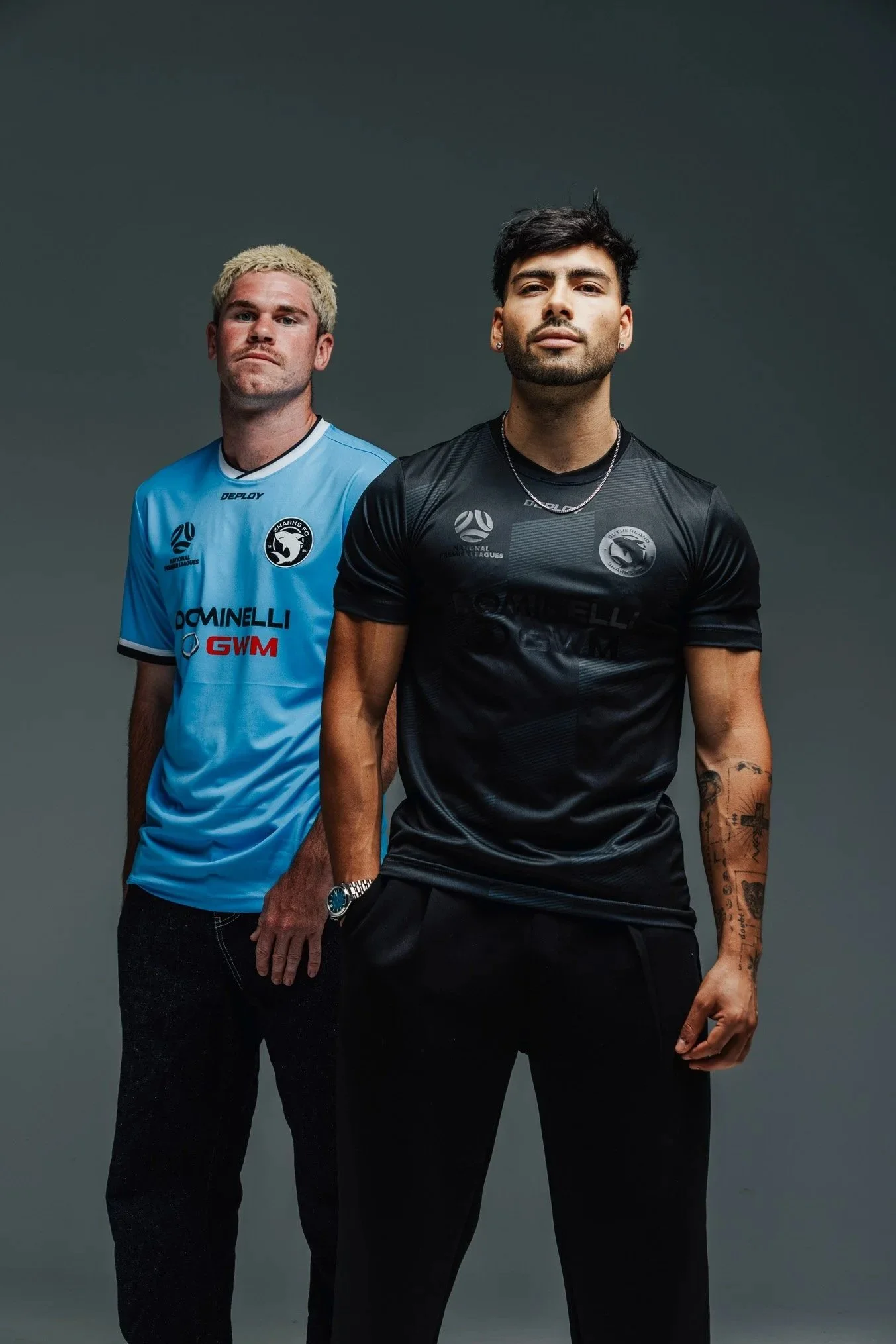 Two men standing in front of a gray background, wearing sports jerseys with logos, one in blue and the other in black, looking at the camera.