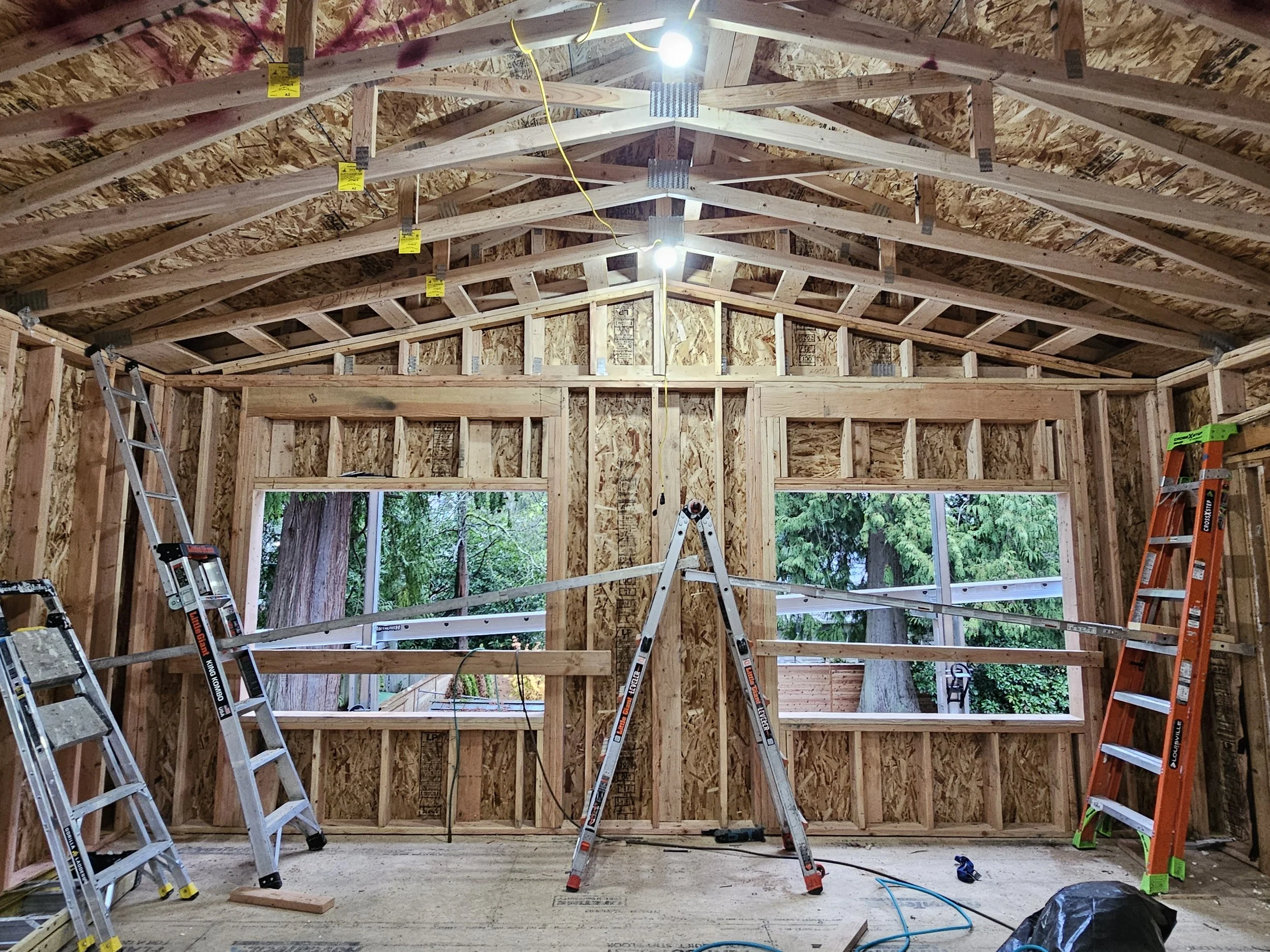 Interior of a house under construction with exposed wooden framing, two large windows, and three ladders set up inside. Construction lights are hanging from the ceiling.