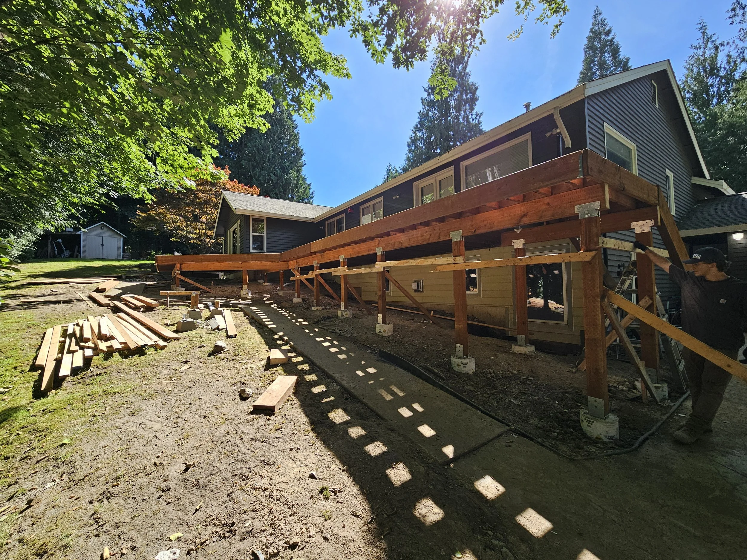 Construction workers building a wooden deck at a residential house, with tools and wood planks on the ground, surrounded by green trees and a clear blue sky.
